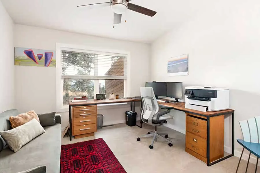 Home office with a desk, computer monitors, and a printer, a white swivel chair, a window with blinds, a small sofa with pillows, a red rug, and wall art