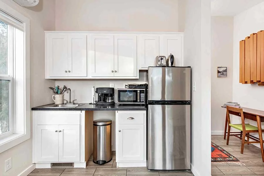 Kitchen with white cabinets, black countertop, stainless steel refrigerator, microwave, coffee maker, toaster, and a window, with a small dining area with a wooden table and chair nearby.