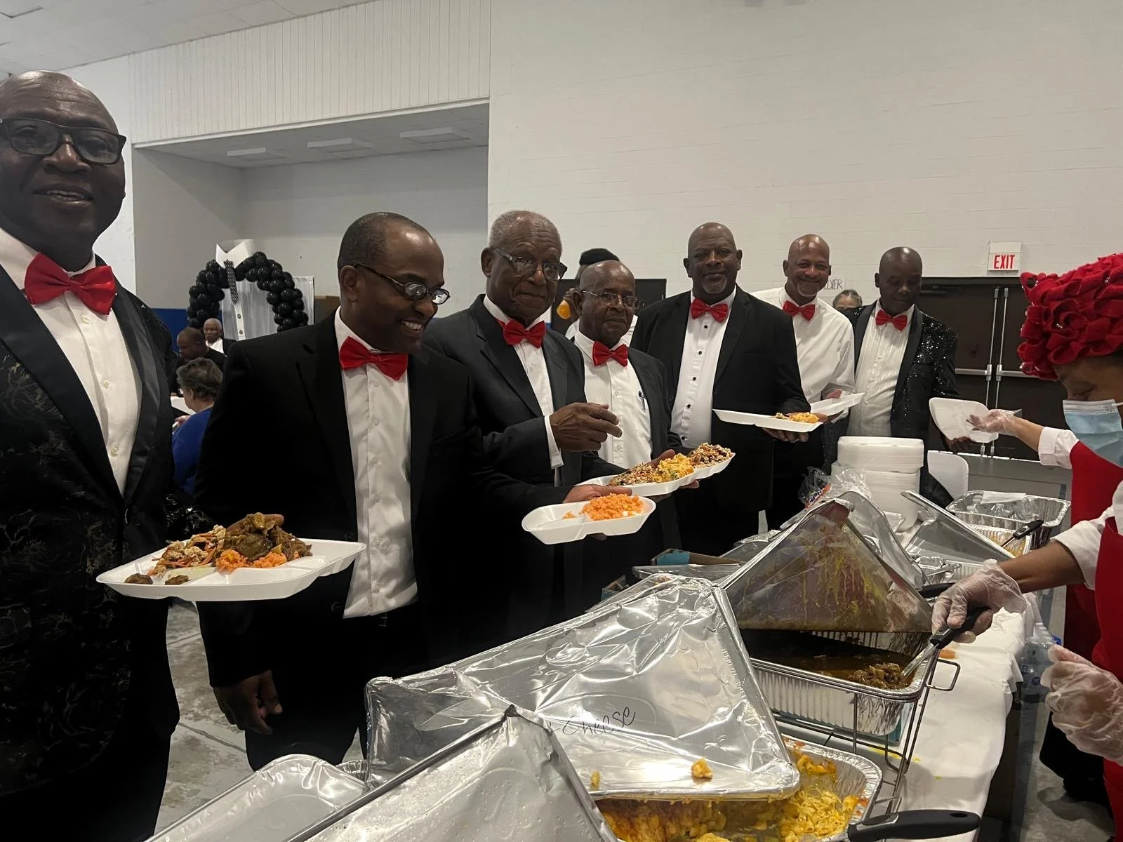 A group of men dressed in tuxedos and red bow ties lining up at a buffet table, holding disposable plates of food, with a woman wearing a red head covering and face mask serving food.