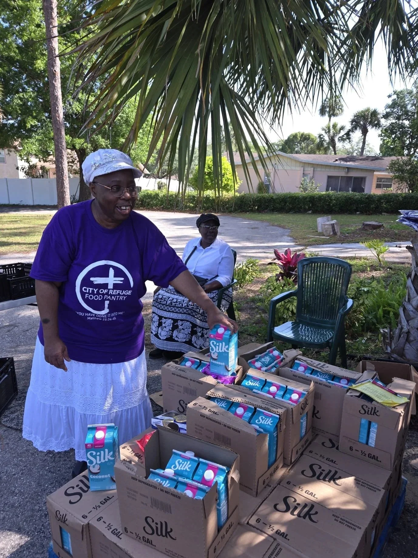 Two women, one elderly woman and one middle-aged woman, standing outside near boxes of silk coconut milk cartons, with trees and a house in the background, under a large palm tree. The elderly woman is wearing a purple shirt with text and a white ski