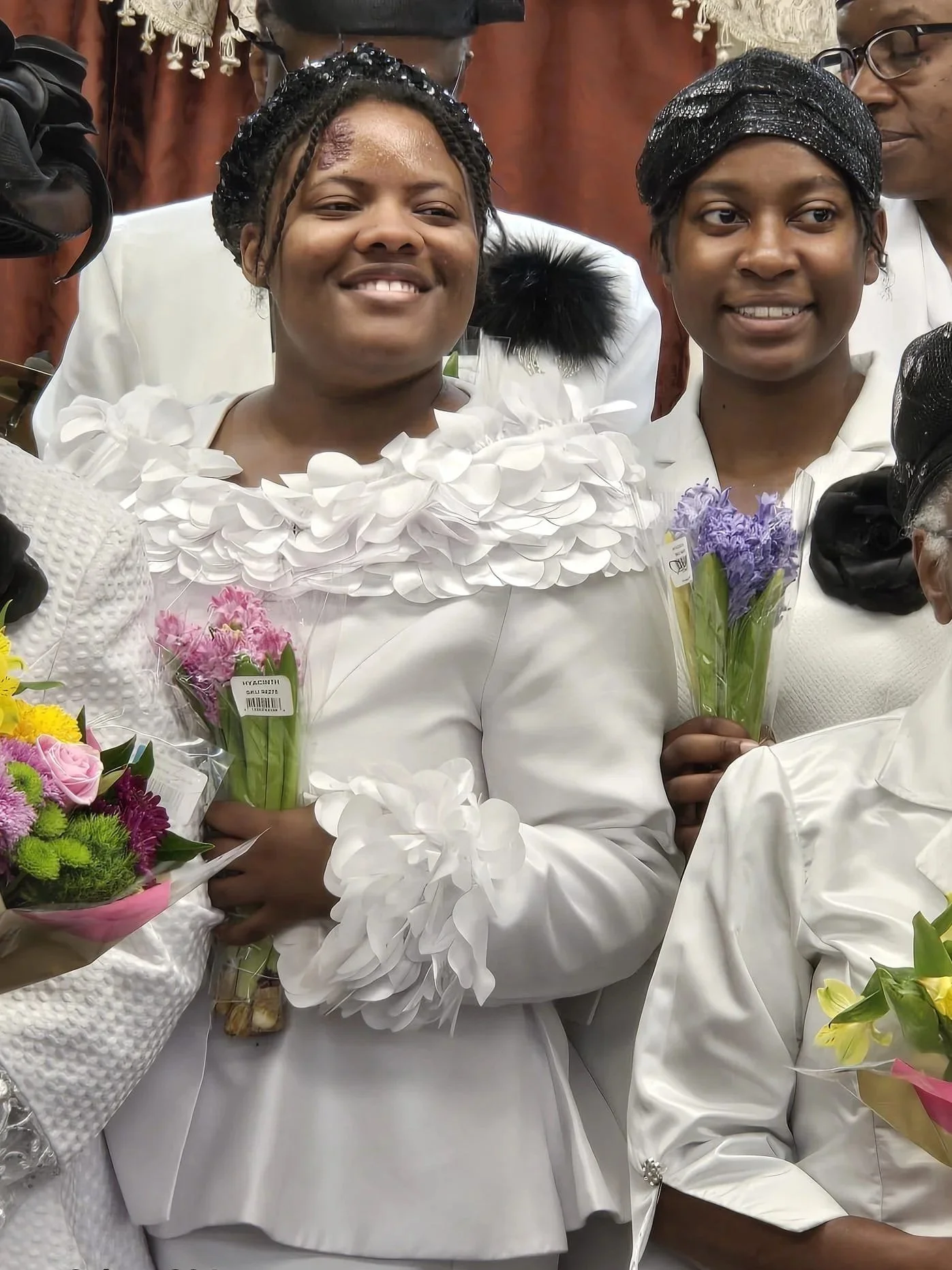 Two women dressed in white holding bouquets of pink and purple flowers, smiling in a celebratory setting.