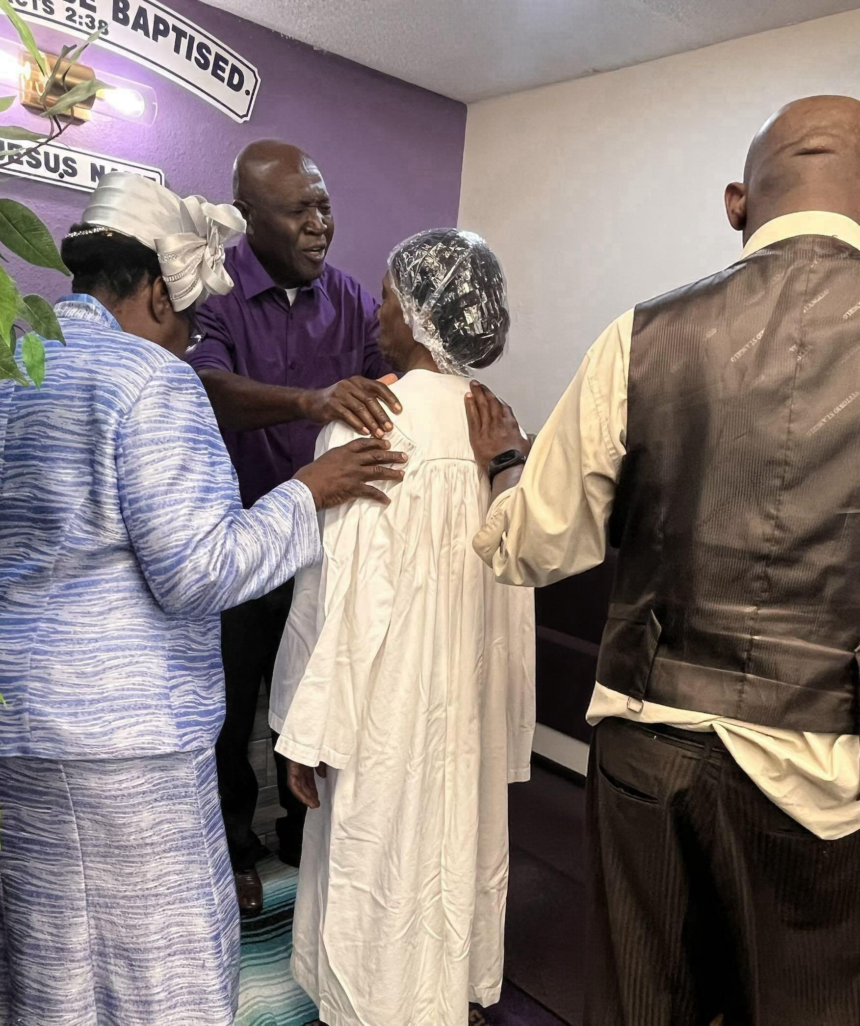 A group of four individuals are gathered closely together, engaged in a discussion or prayer. They are in a room with a purple and white wall, with religious signs visible in the background, including a sign that says 'Baptized' and another that says