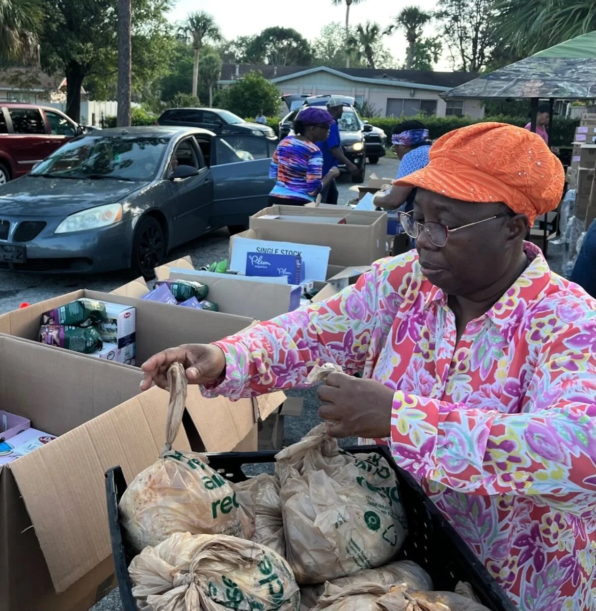 A woman in a colorful floral shirt and orange headscarf is packing groceries into paper bags at an outdoor distribution event. Several cardboard boxes and parked cars are visible in the background, with other women in the distance.