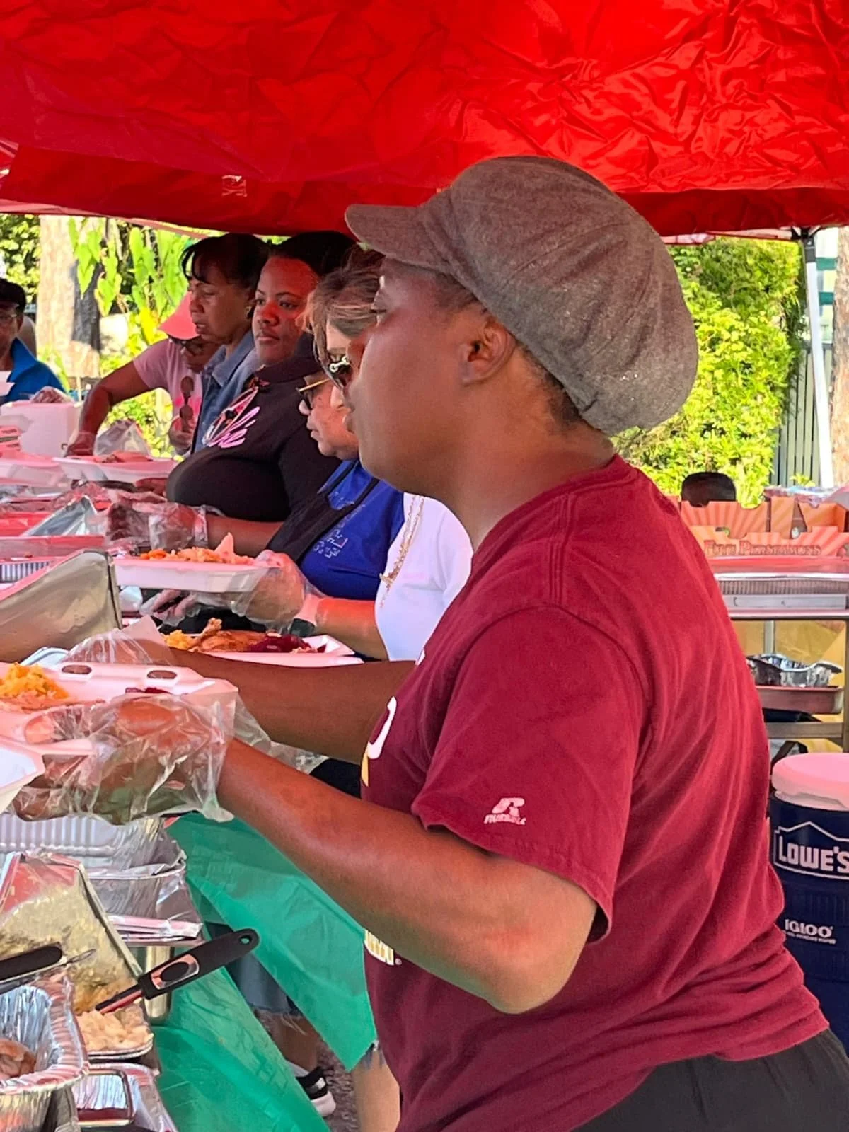 A group of diverse women standing at a buffet table outdoors under a red canopy, serving food and wearing casual clothing.