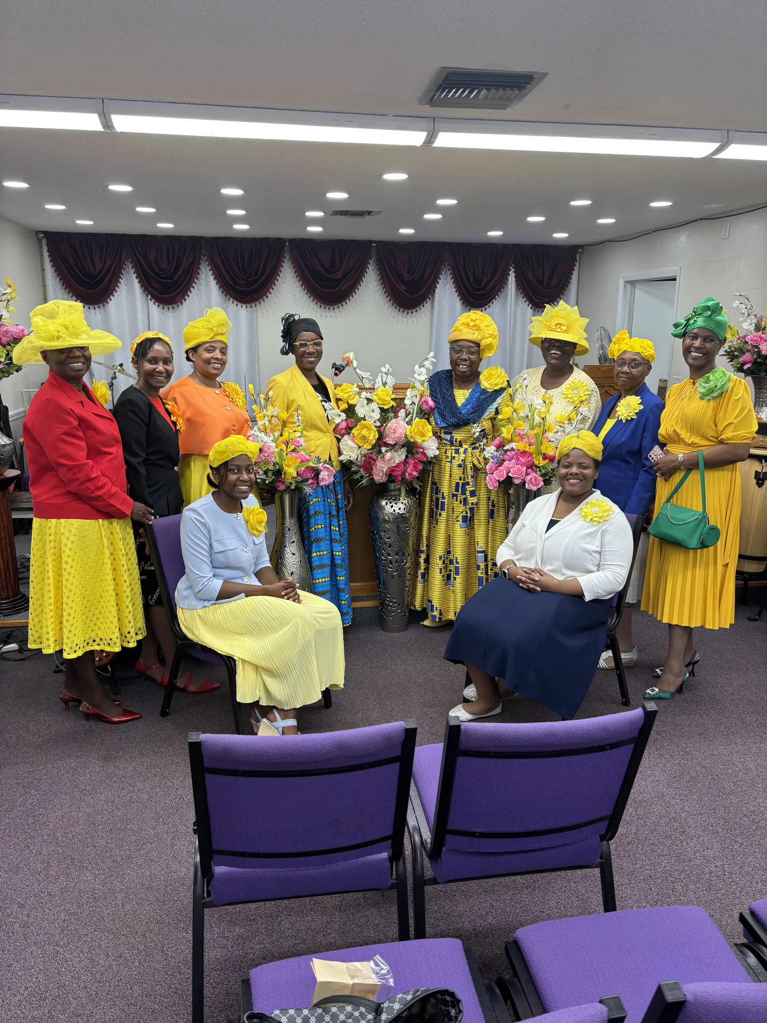 Ten women dressed in colorful traditional attire, including yellow, red, blue, white, and black outfits, gathered indoors for a group photo. They are smiling and standing behind floral arrangements in vases, with purple curtains and white drapes in t