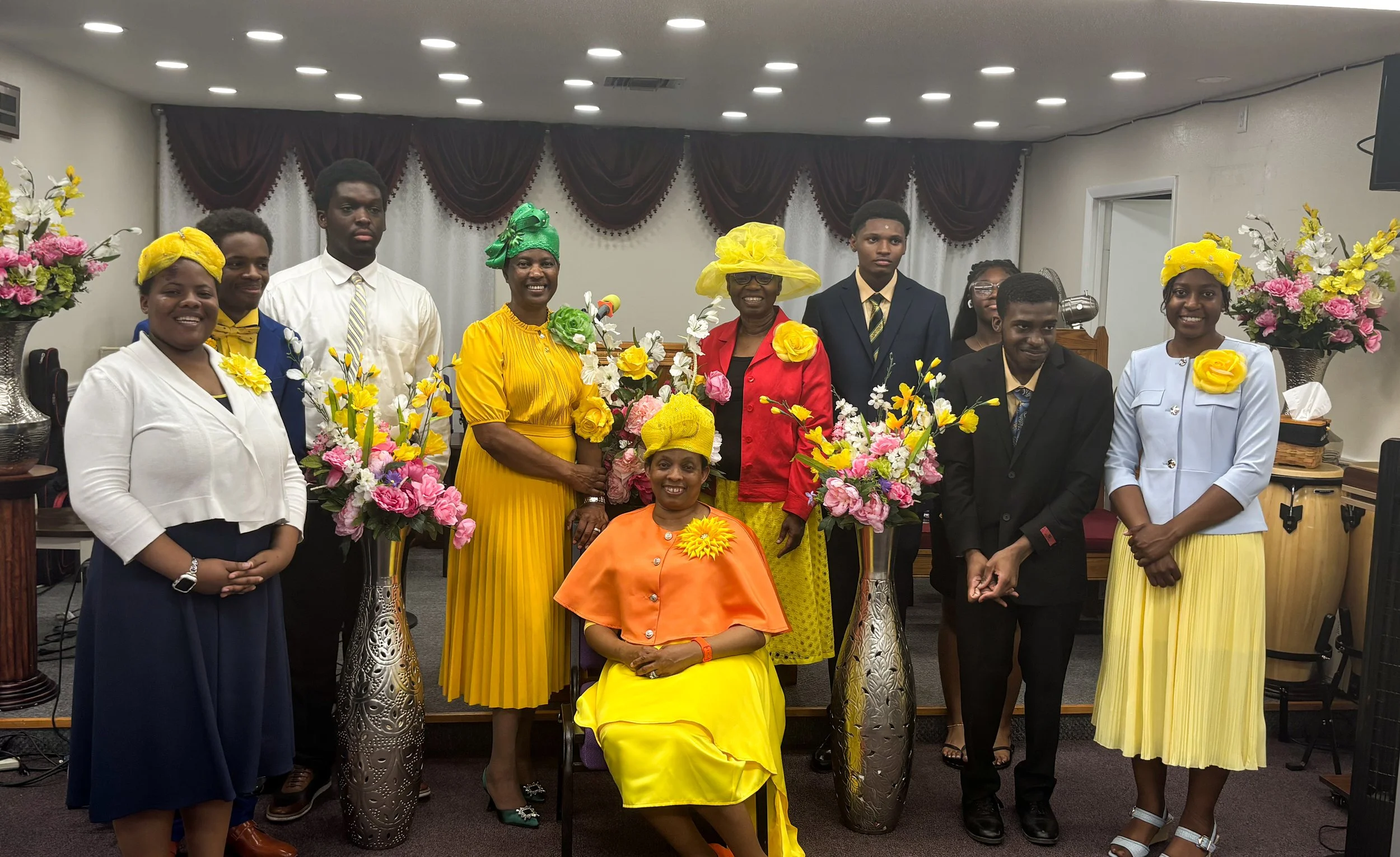 Group of people standing and sitting in a room decorated with flowers, with women wearing yellow, green, and white outfits and men in suits, during a celebratory event.