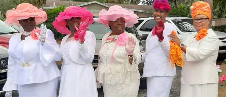 Five women dressed in white outfits and colorful hats, standing outdoors in front of parked cars.