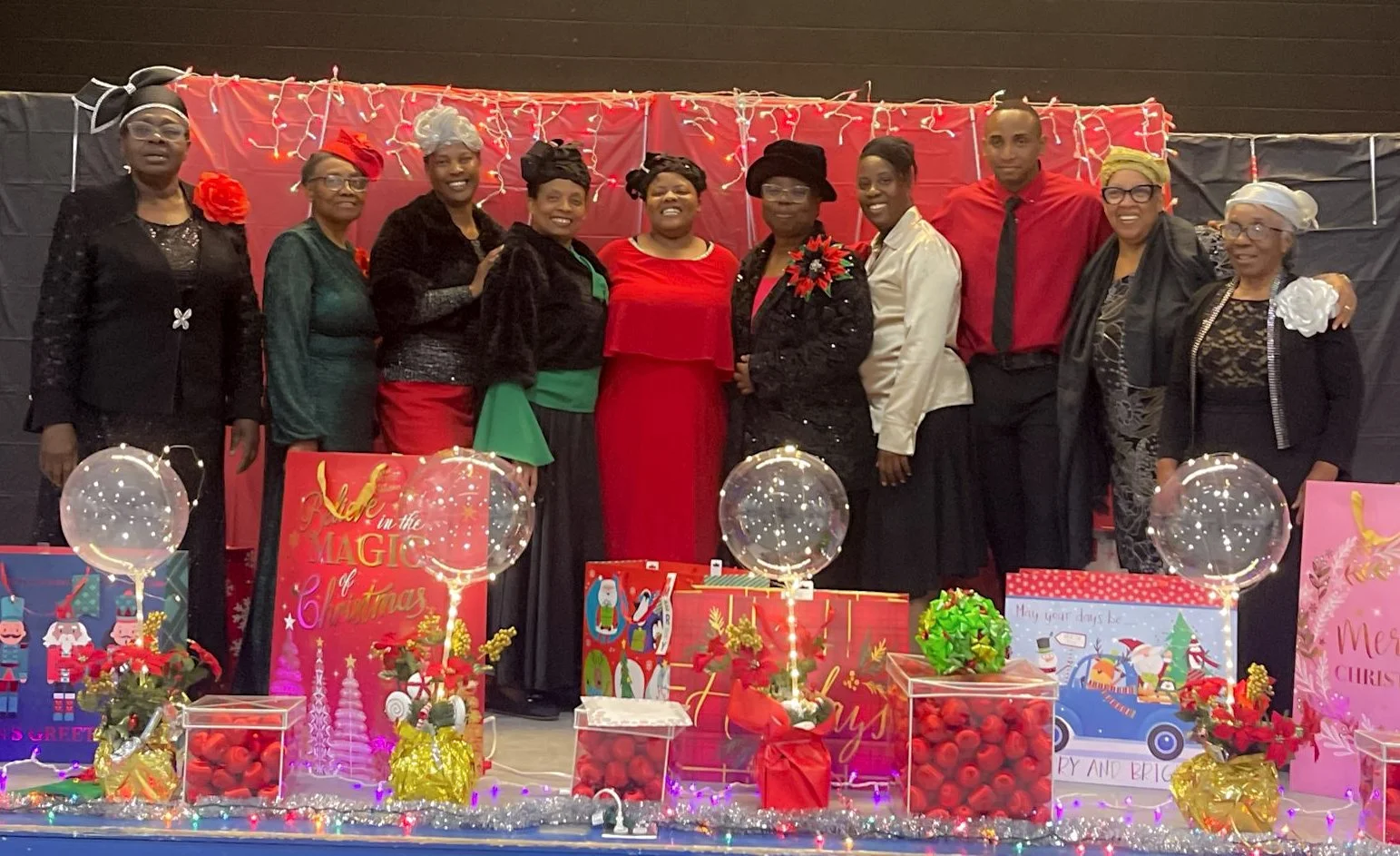 A group of eleven people dressed in festive attire, standing on a decorated stage with Christmas-themed decorations, including wrapped presents, balloons, and lights.