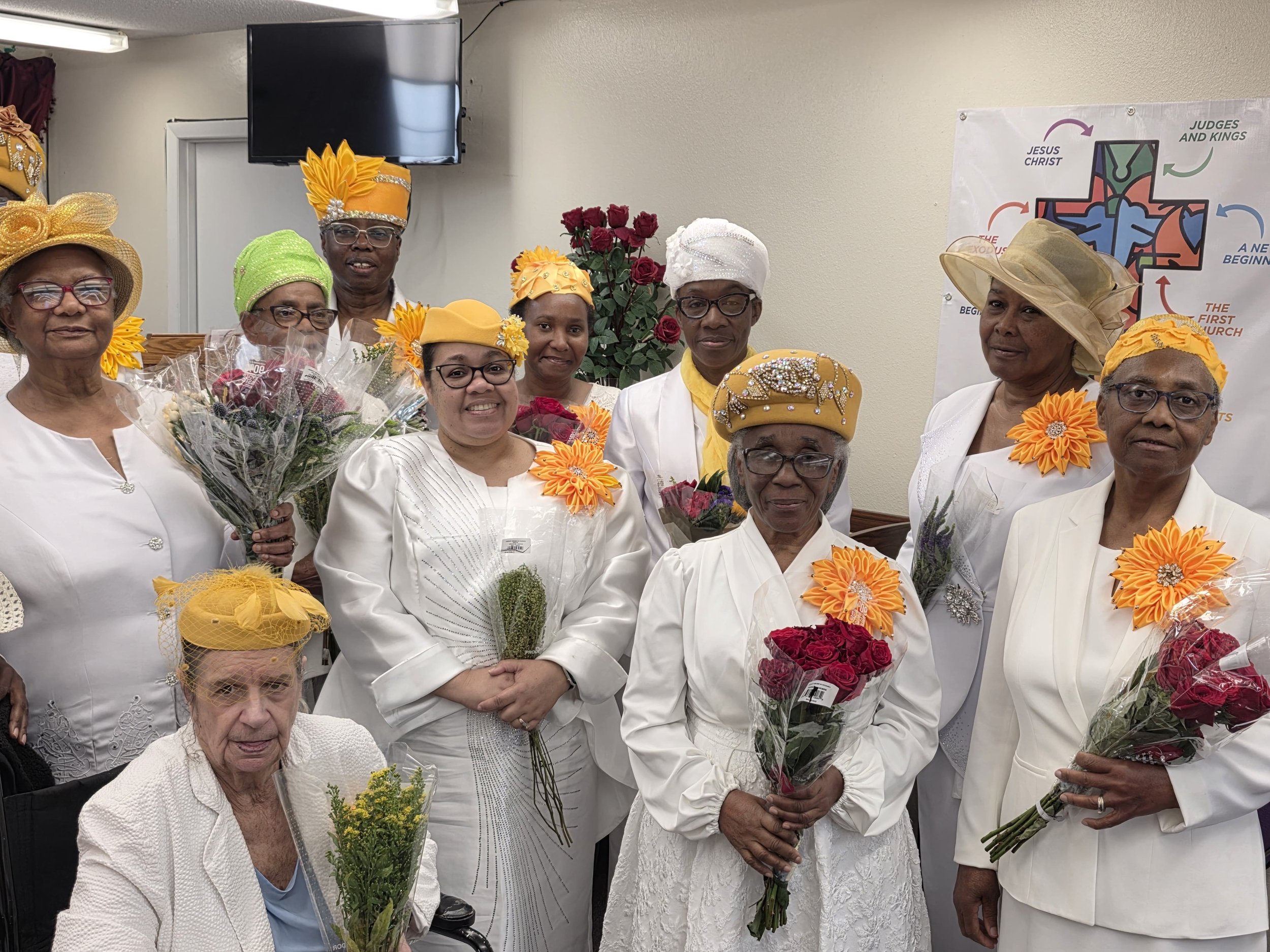 Group of elderly women dressed in white, wearing colorful hats and holding bouquets of flowers, in a church or community center setting.