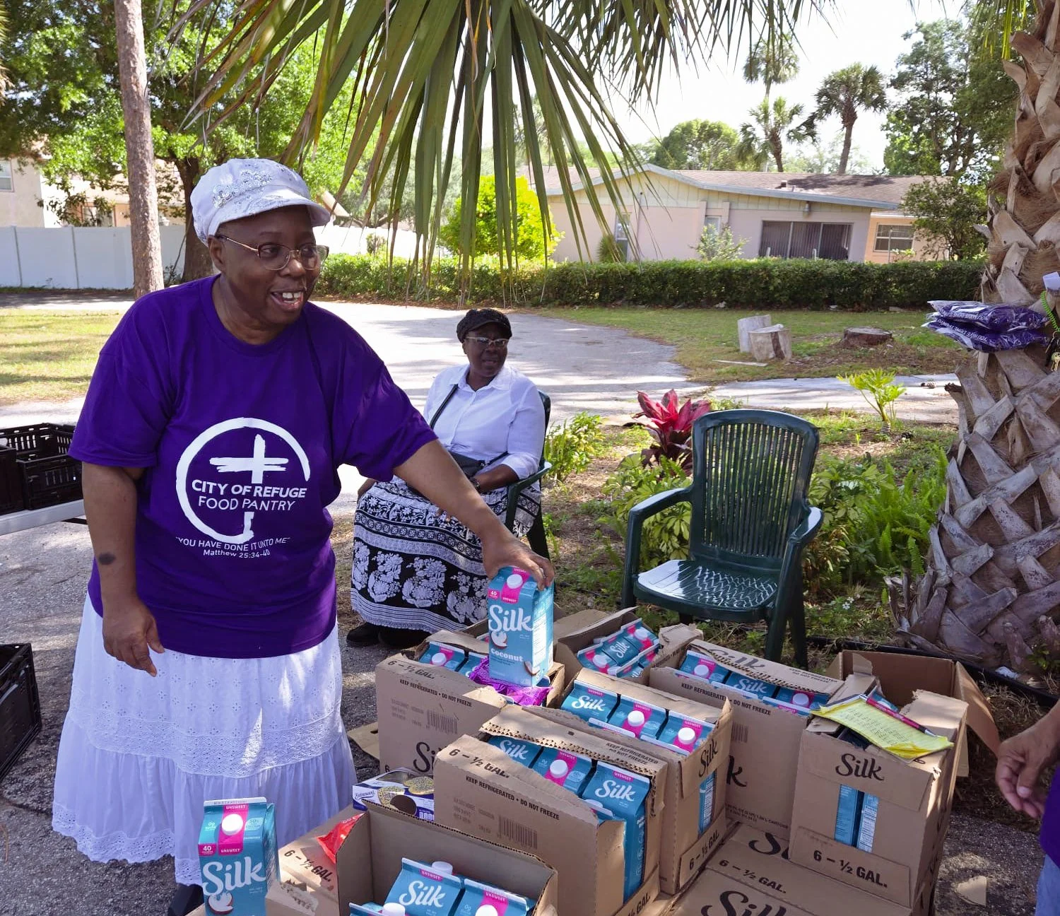 Two women at an outdoor food pantry distributing cartons of Silk coconut milk. One woman, wearing a purple shirt with logo of City of Refuge Food Pantry, is reaching for a carton, while the other woman, wearing a white blouse and skirt, is seated behind the table surrounded by boxes of Silk milk.