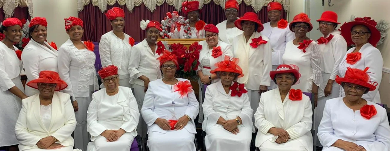 Group of women dressed in white, wearing red hats and accessories, gathered indoors in celebration.