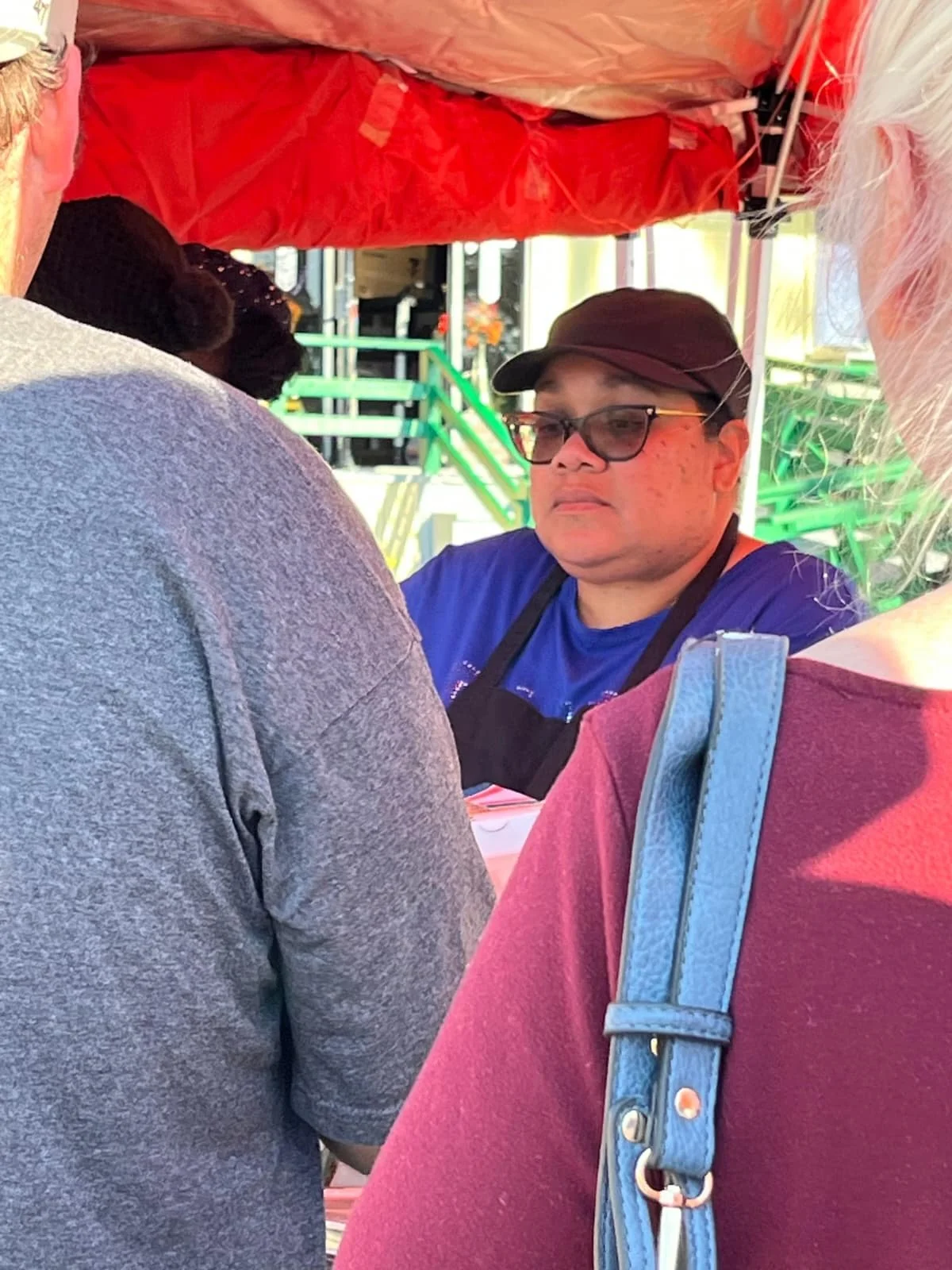 A man wearing glasses, a black cap, and a blue shirt stands behind a food stall. Several people are around, and the background shows green stairs and bright lights.