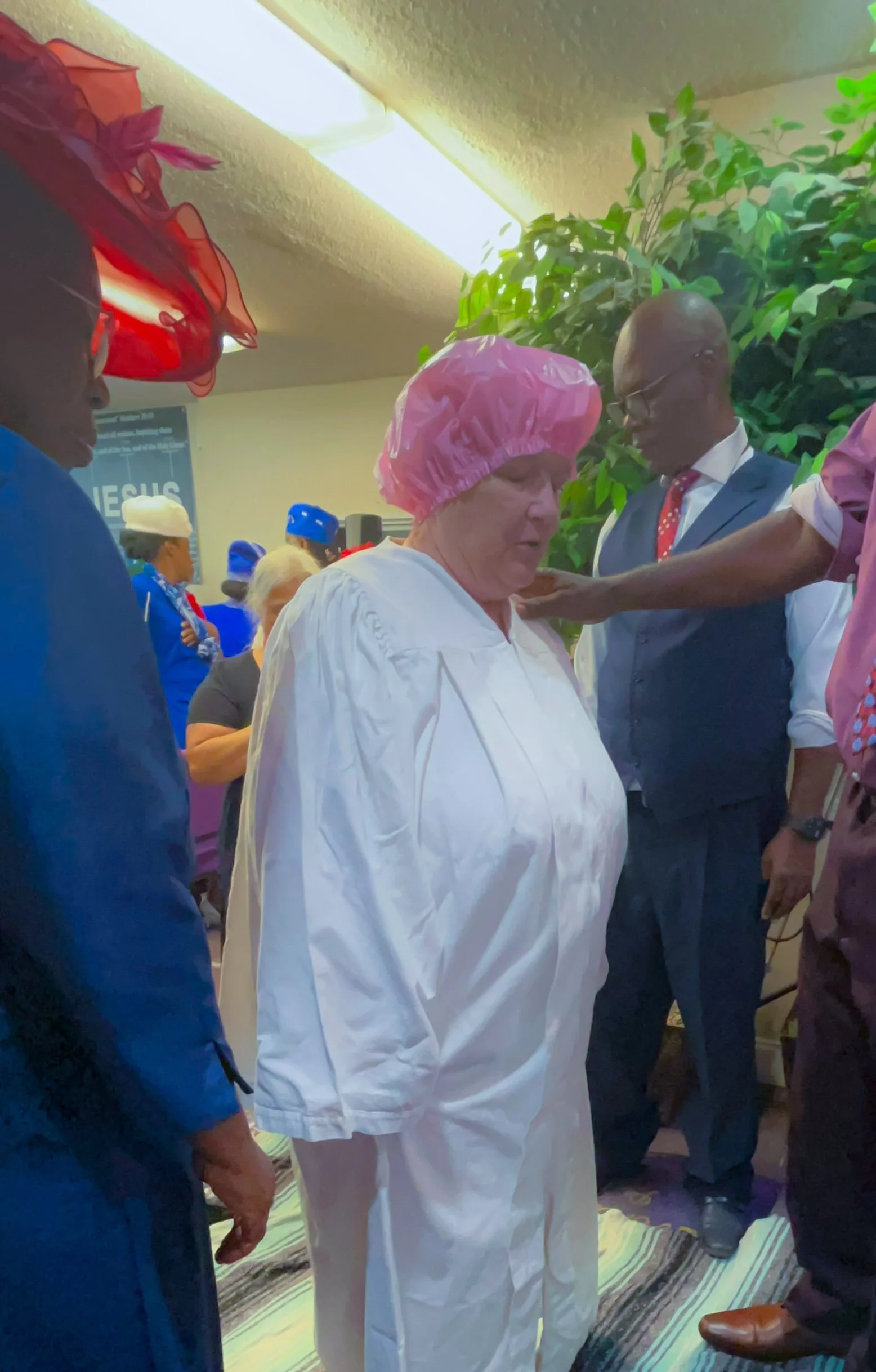 Group of people engaged in a prayer or spiritual ceremony indoors, with some wearing head coverings, including a woman in a pink shower cap and a man in a suit, near a green leafy plant.