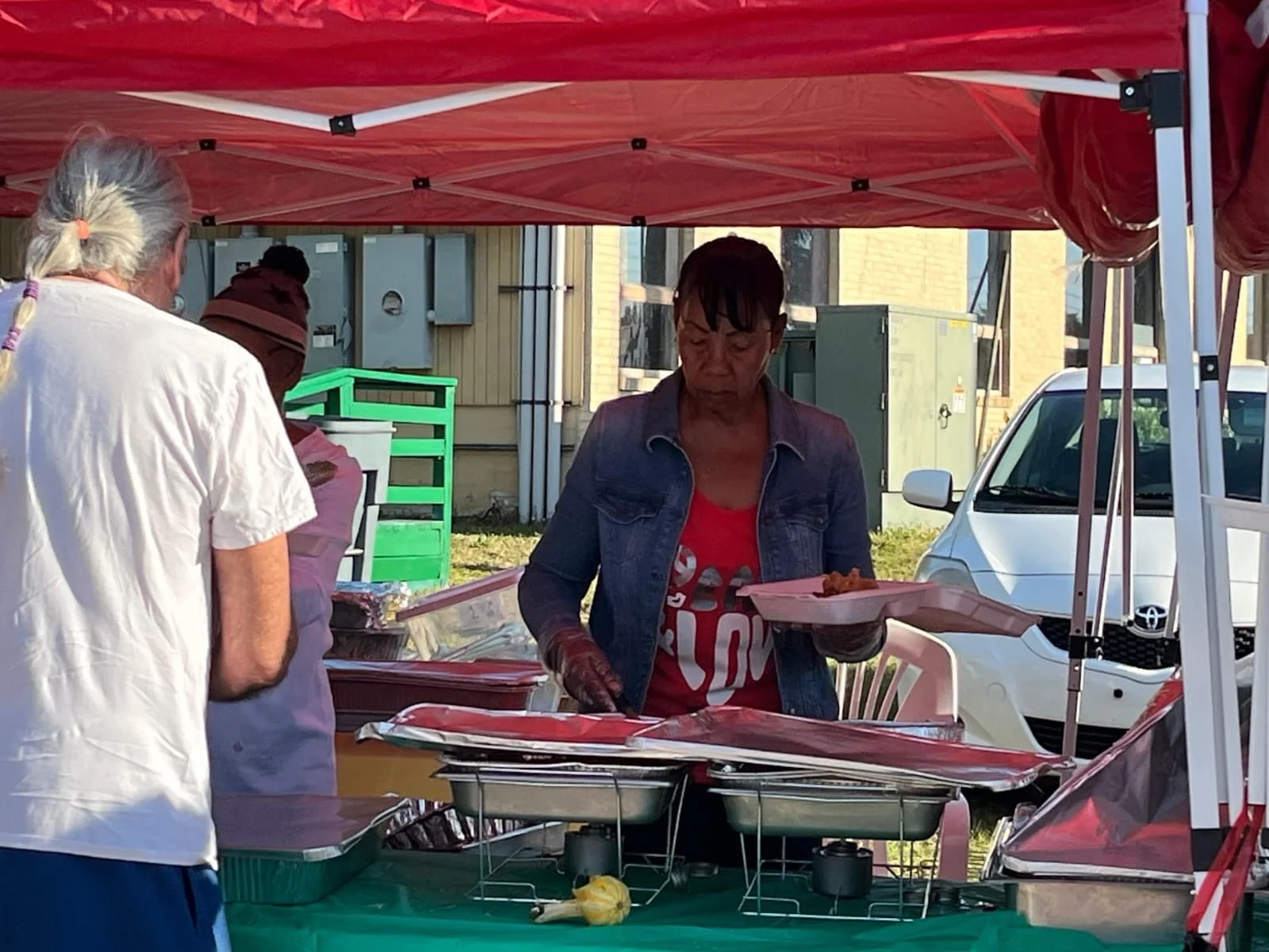 A woman serving food at an outdoor market stall with a red canopy, with several people, cars, and utility boxes in the background.