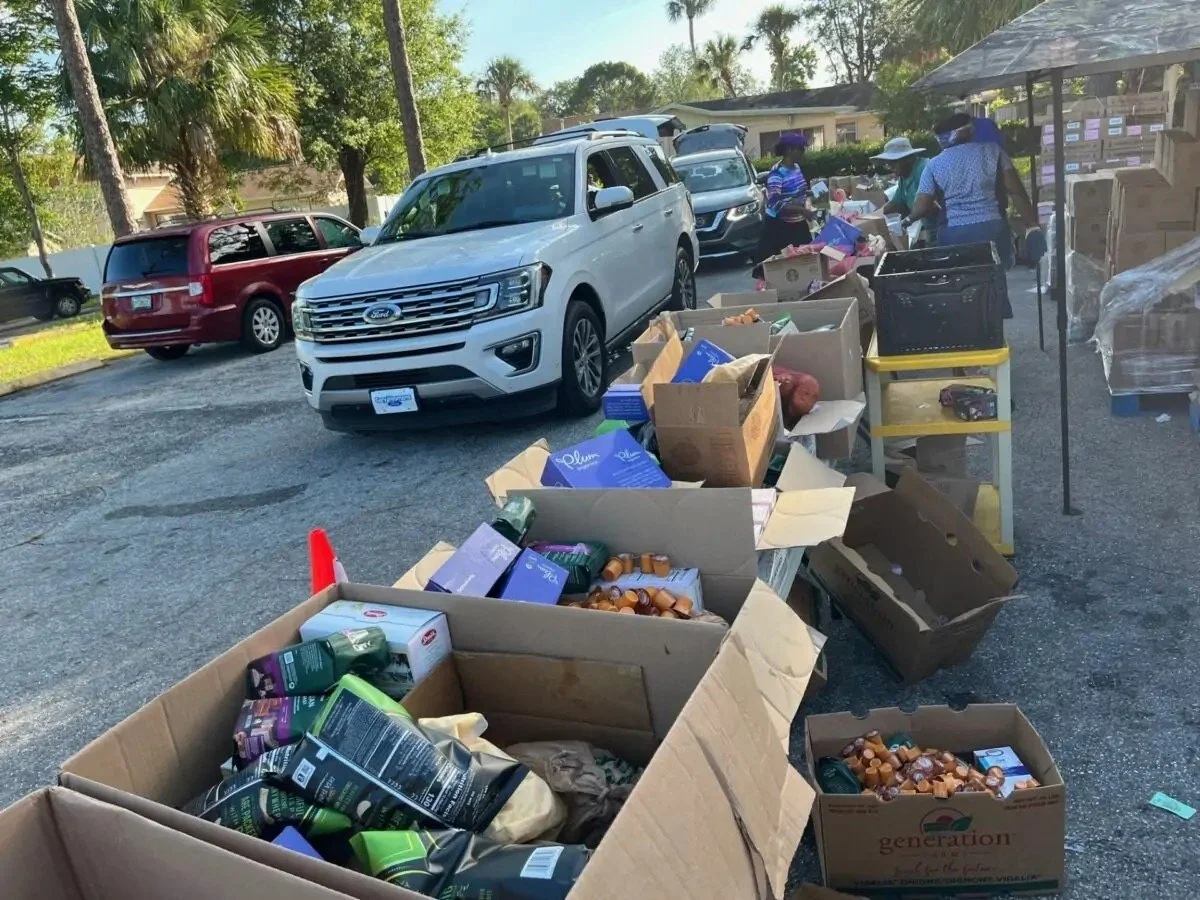 People shopping outdoors with cars parked in background, and boxes of various products on tables and the ground.