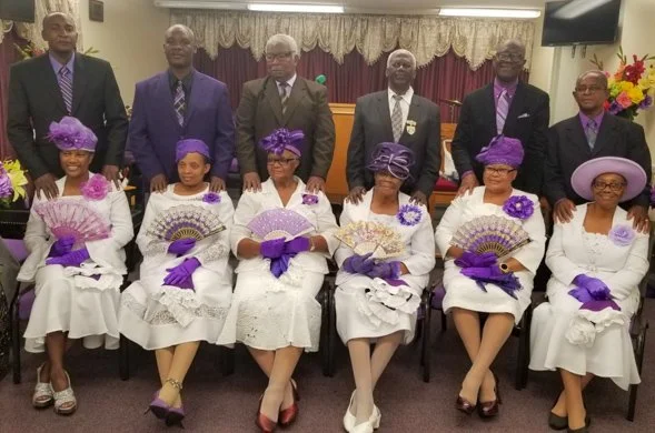 Group of seven women seated in front, dressed in white dresses with purple accessories and fans, and six men standing behind them in formal suits, in a decorated room with floral arrangements and curtains.