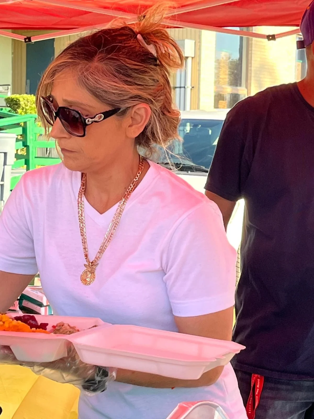 Woman with short reddish hair, wearing glasses, gold jewelry, and a white T-shirt, holding a pink food tray at an outdoor event.