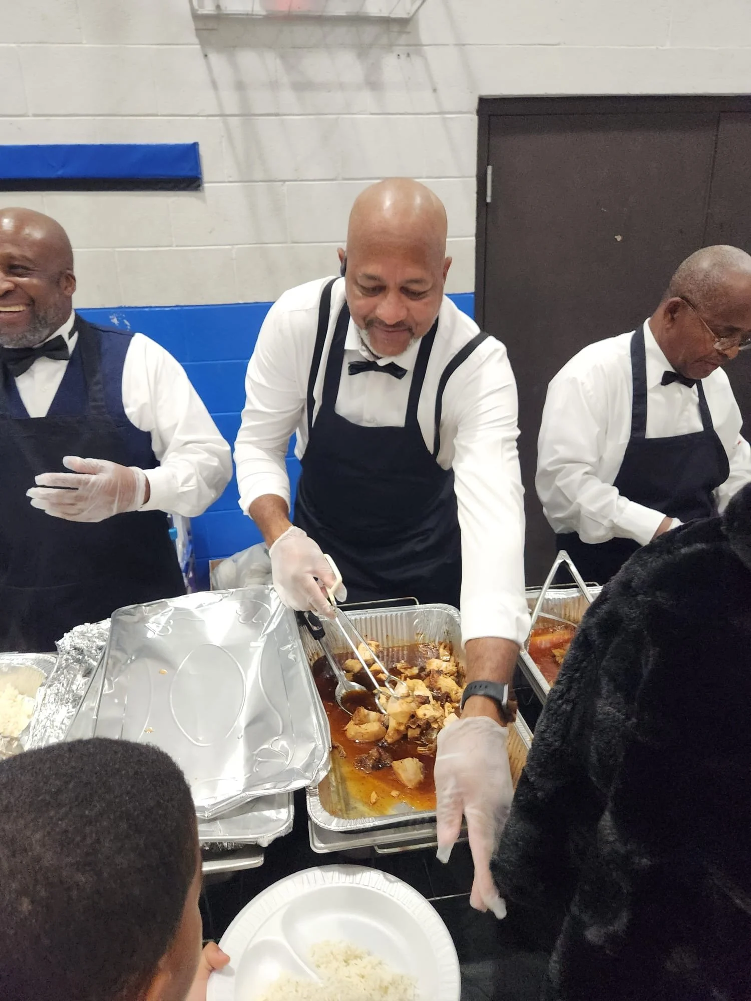 A group of men dressed in formal attire with aprons and gloves serving food at a buffet table, with trays of cooked dishes and a bowl of rice in the foreground.