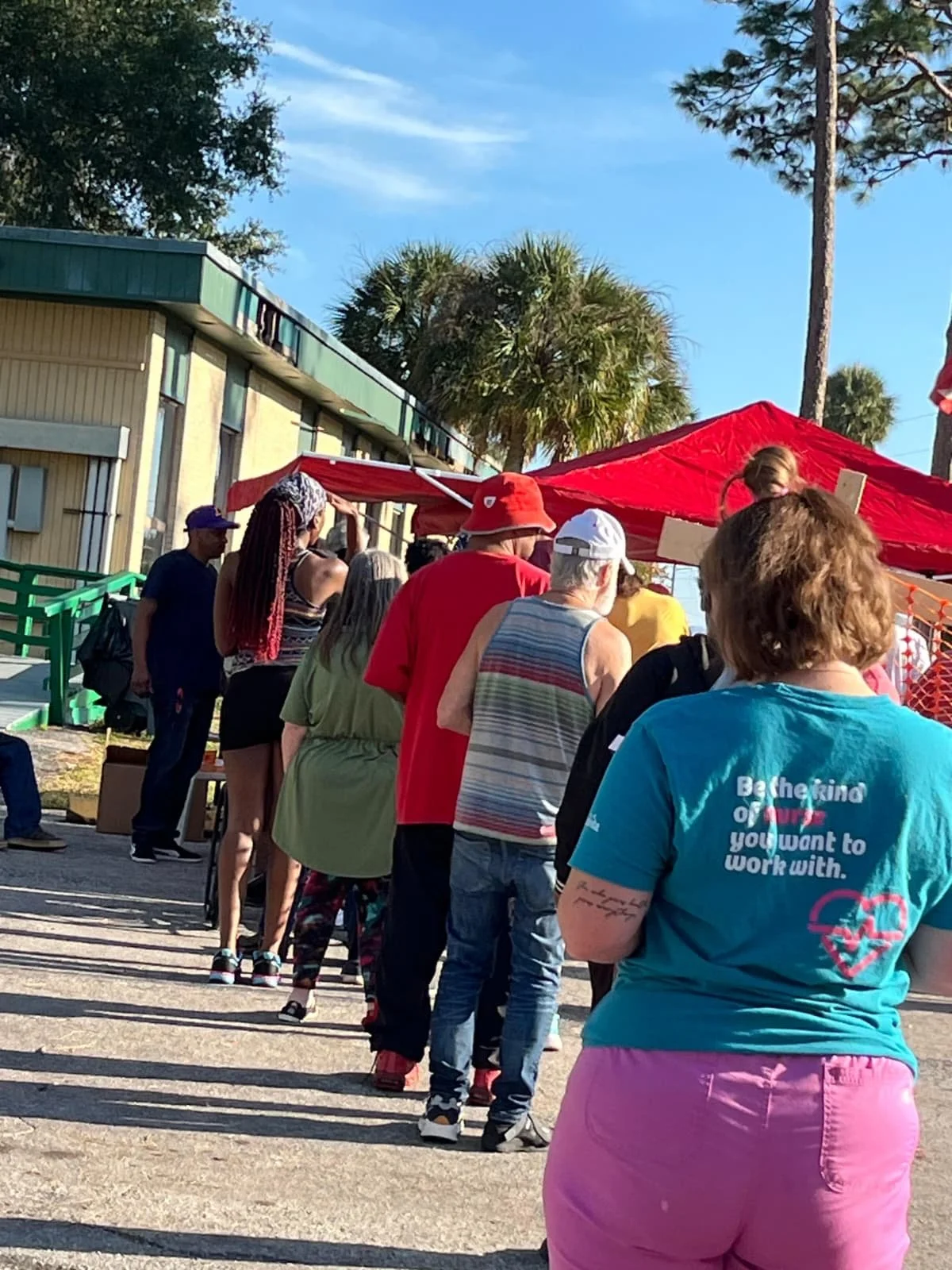 People waiting in line outdoors at a community event under a red canopy with palm trees in the background on a sunny day.