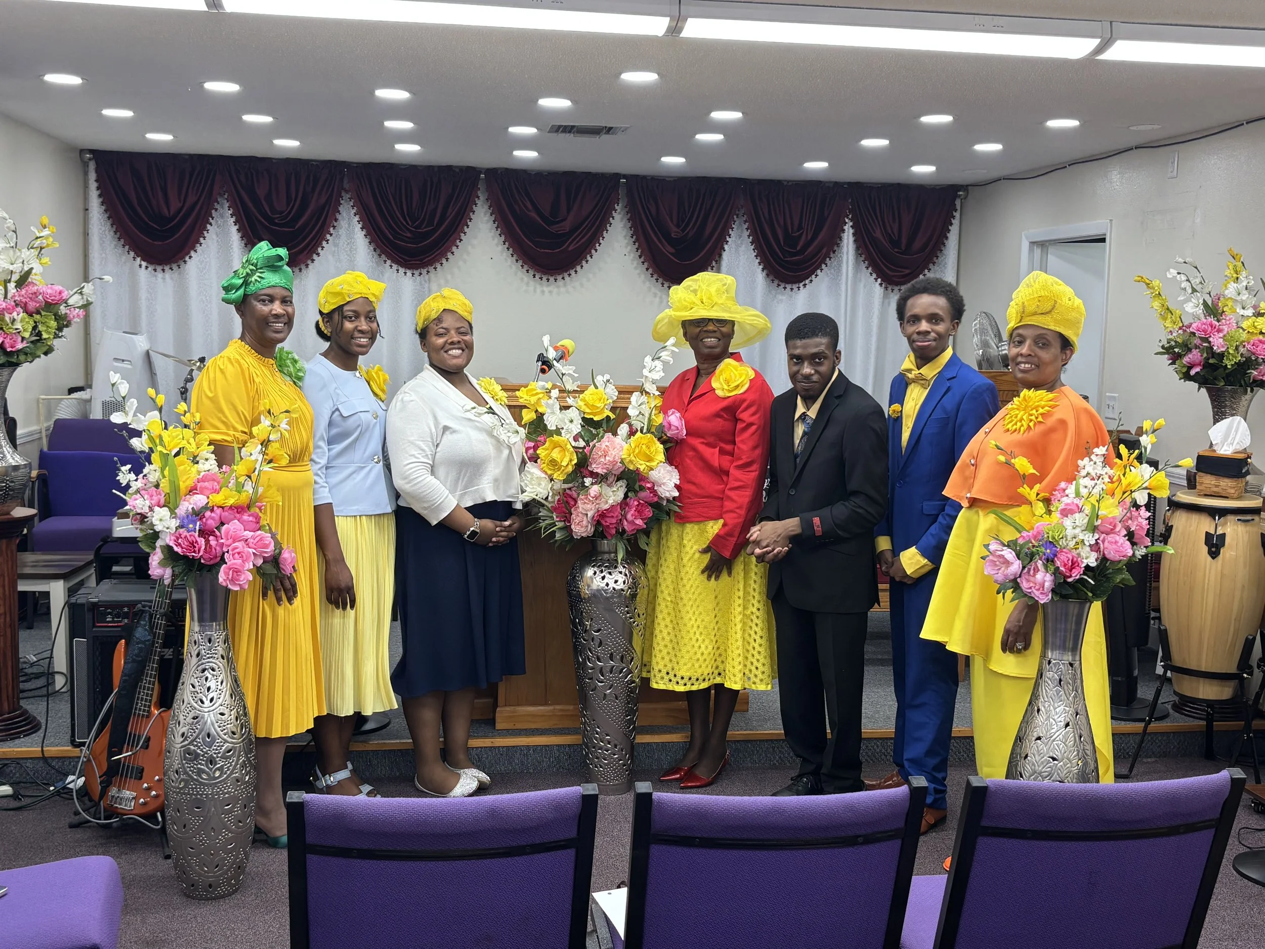 A group of seven people dressed in colorful clothing posed together indoors, surrounded by flower arrangements and standing in front of a dark red curtain with scalloped edges.