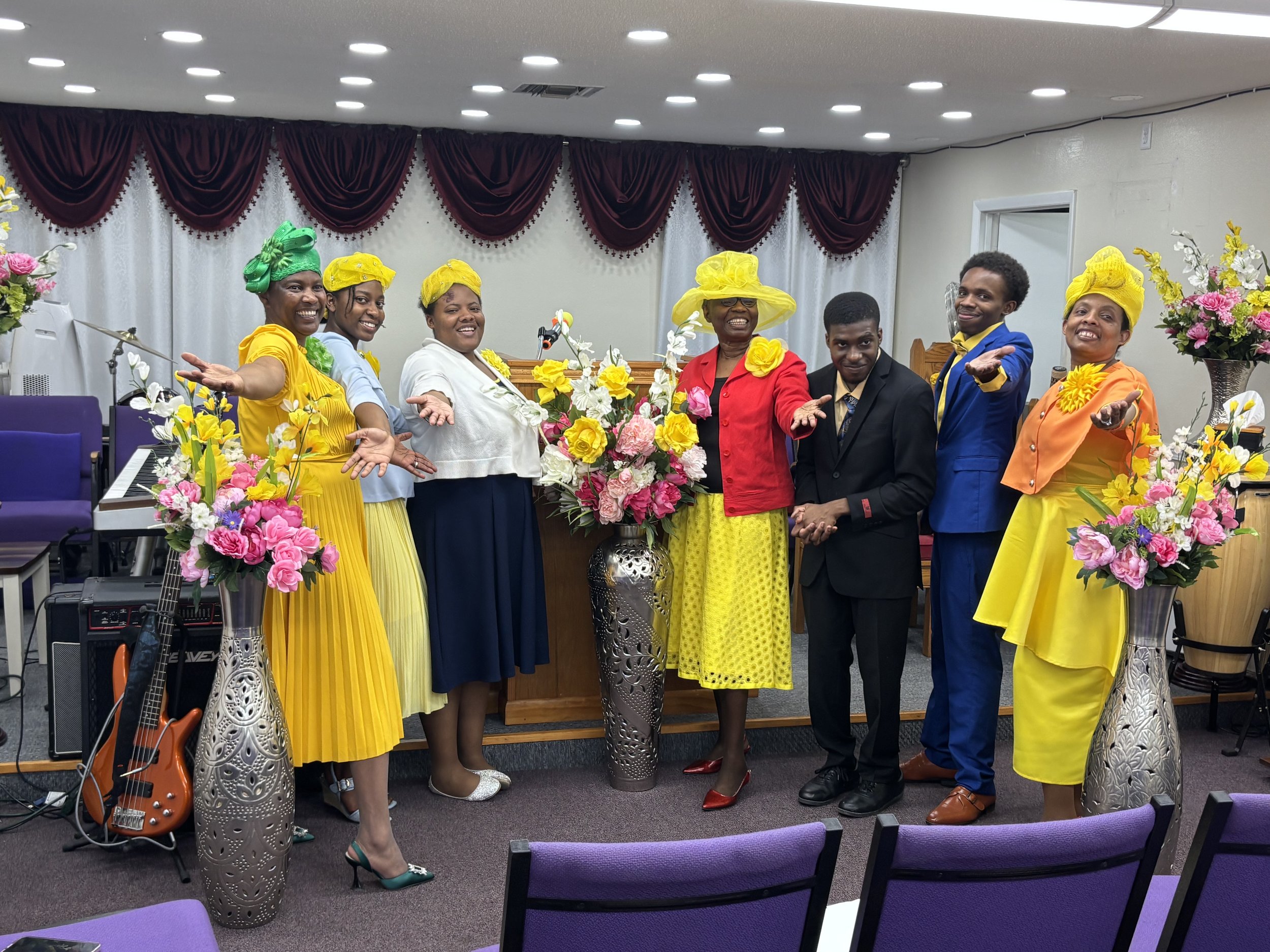 Group of six people dressed in colorful formal attire standing in front of a flower arrangement in a decorated room. The women wear bright dresses and hats, and the men wear suits. They are smiling and posing with open arms.