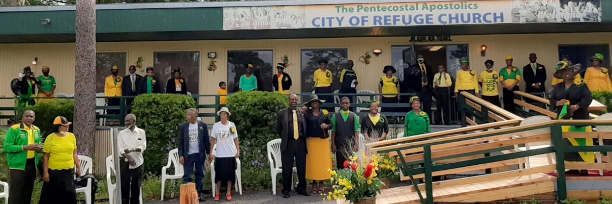 People gathered in front of City of Refuge Church for a community event. Some are in bright yellow and green shirts and jackets, and others are dressed in formal attire. A flower arrangement is on the ground. The church sign reads 'The Pentecostal Apostolics, City of Refuge Church.'