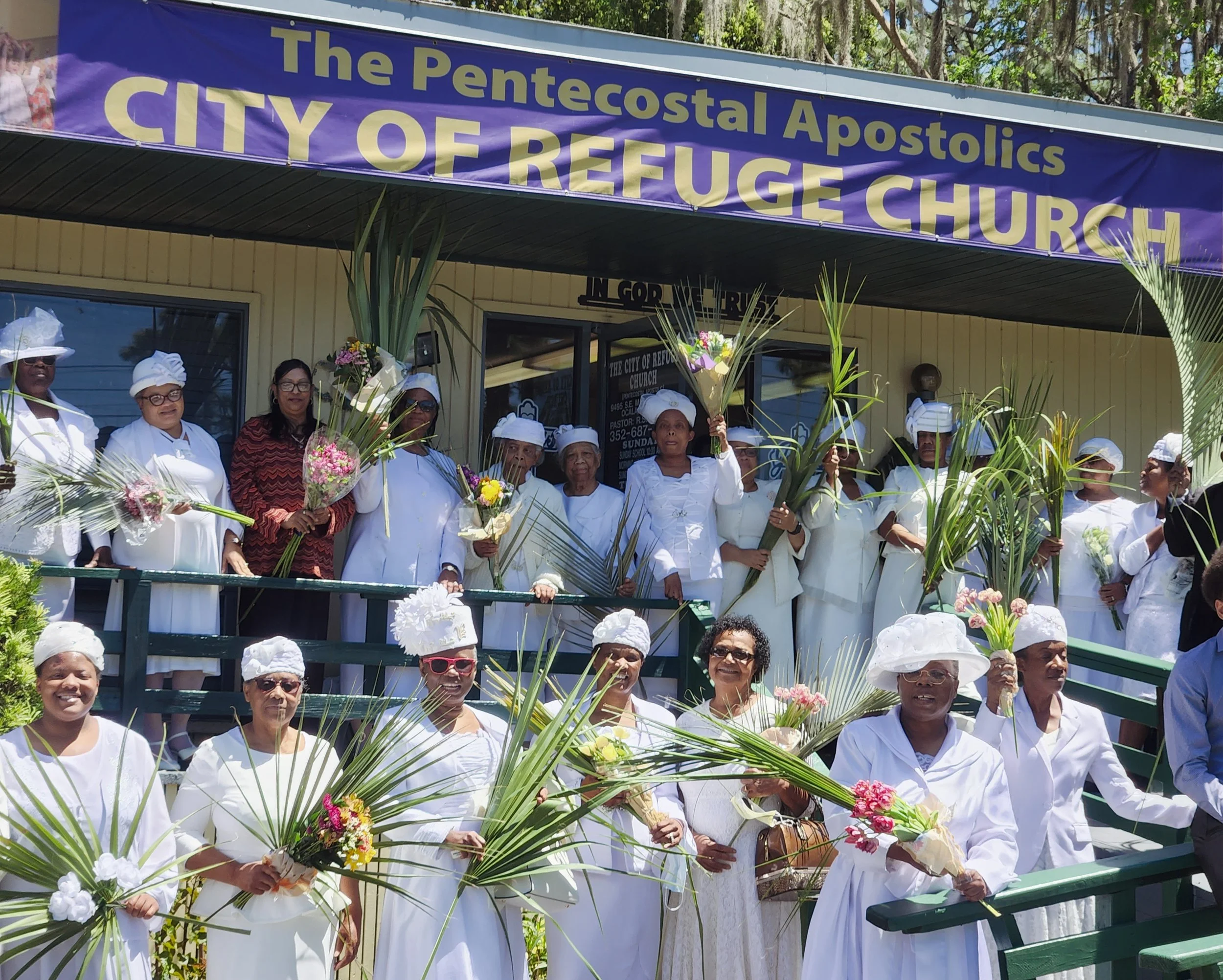 A group of women dressed in white robes and bonnets holding flowers, standing in front of a building with a large purple banner that reads 'The Pentecostal Apostolics CITY OF REFUGE CHURCH'.