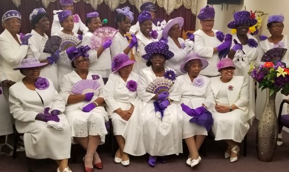 Group of women dressed in white with purple hats, gloves, and accessories, posing for a photo indoors with a flower arrangement nearby.