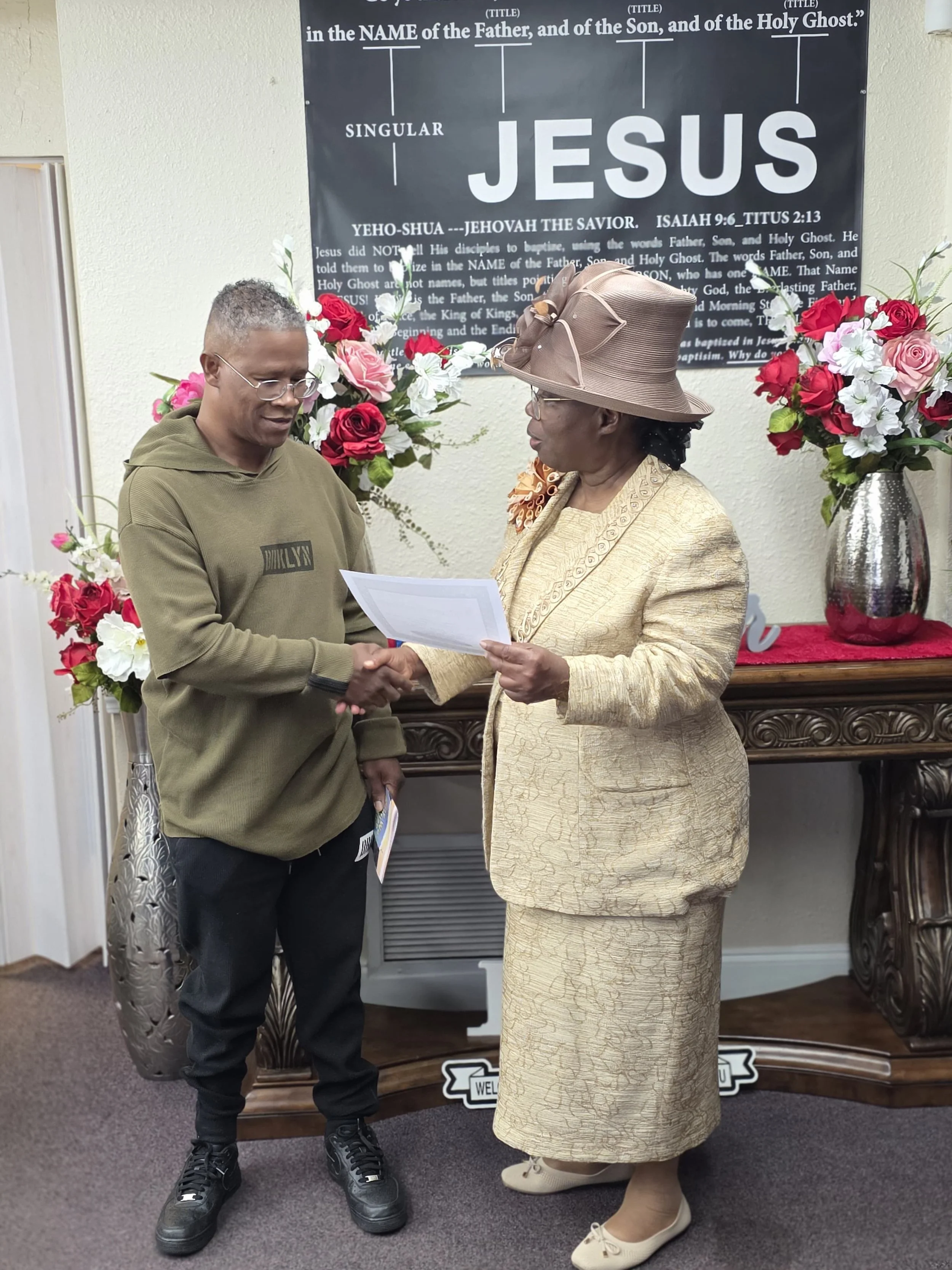 A young man and an elderly woman shaking hands at a religious event, with floral arrangements and a church-themed sign in the background.