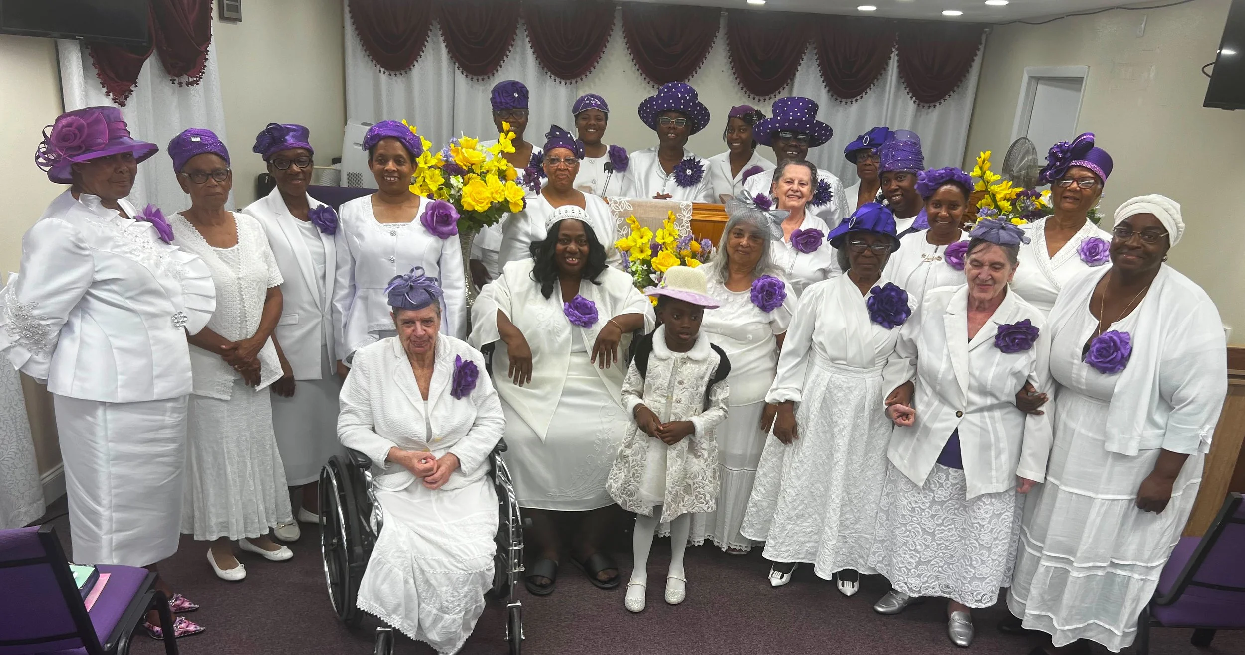 A group of women and girls dressed in white, wearing purple hats and purple roses, gathered for a celebration or special occasion with floral decorations and smiling faces.