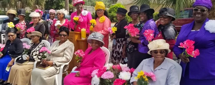 A group of women dressed in colorful hats and flowers, gathered outdoors, some seated and some standing, holding bouquets of pink, yellow, and white flowers.