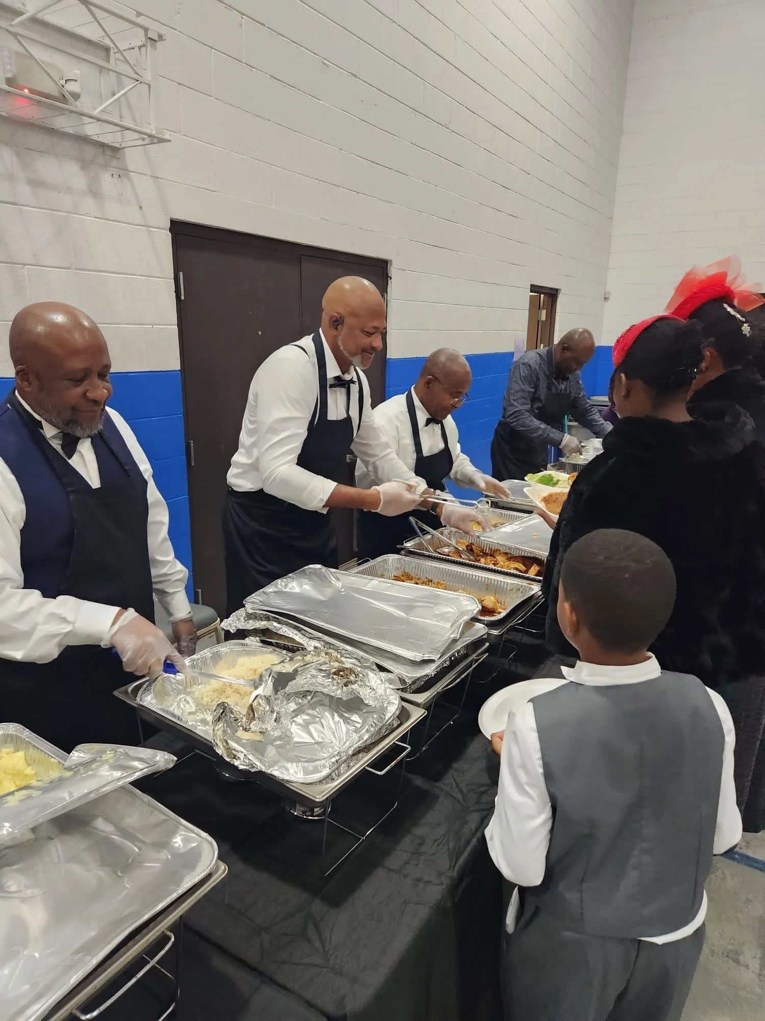 People serving food at a buffet table during a formal event, with children in formal attire watching.
