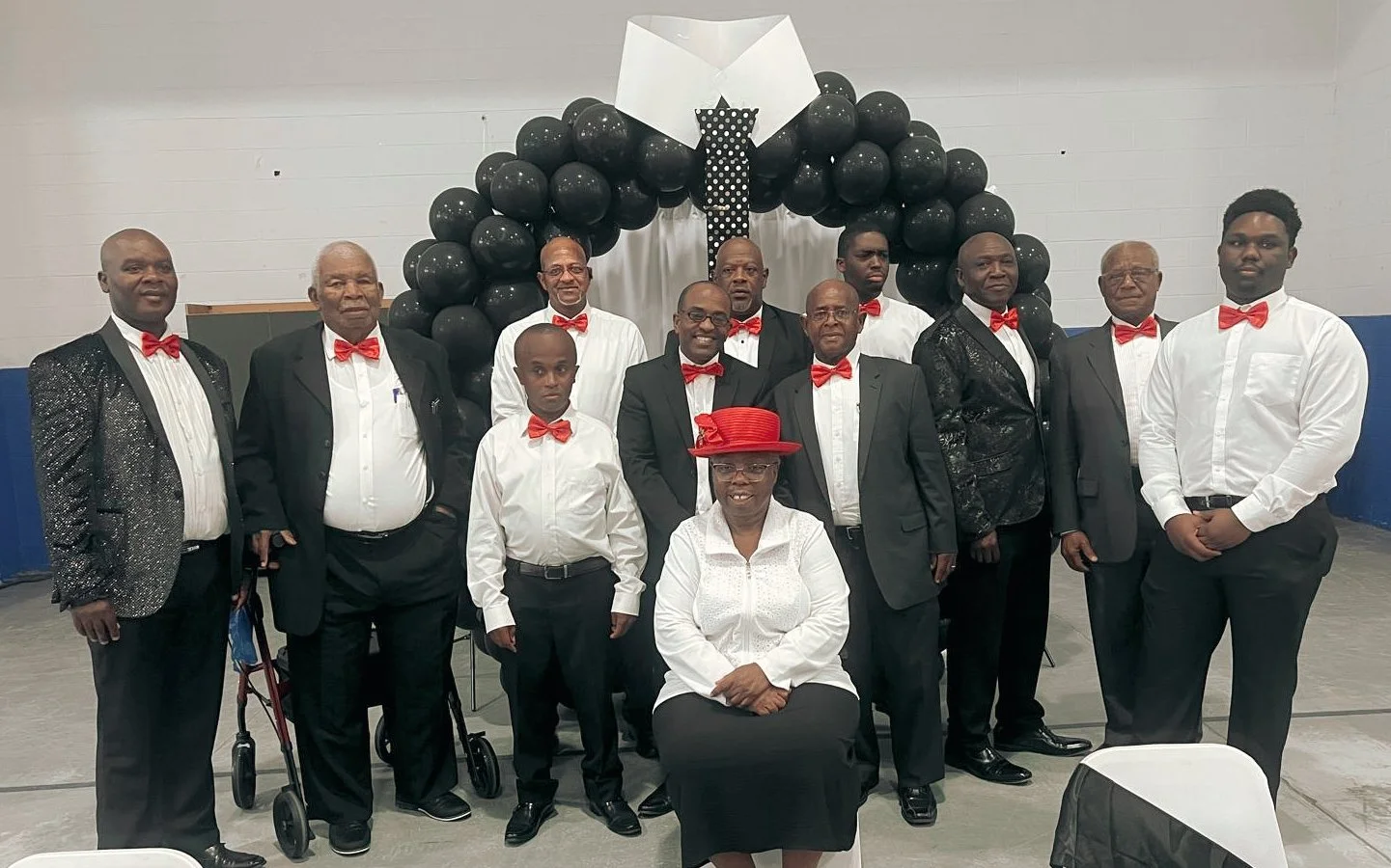 Group of men and one woman dressed in formal attire with red bow ties, posing in front of black balloon arch at indoor event.