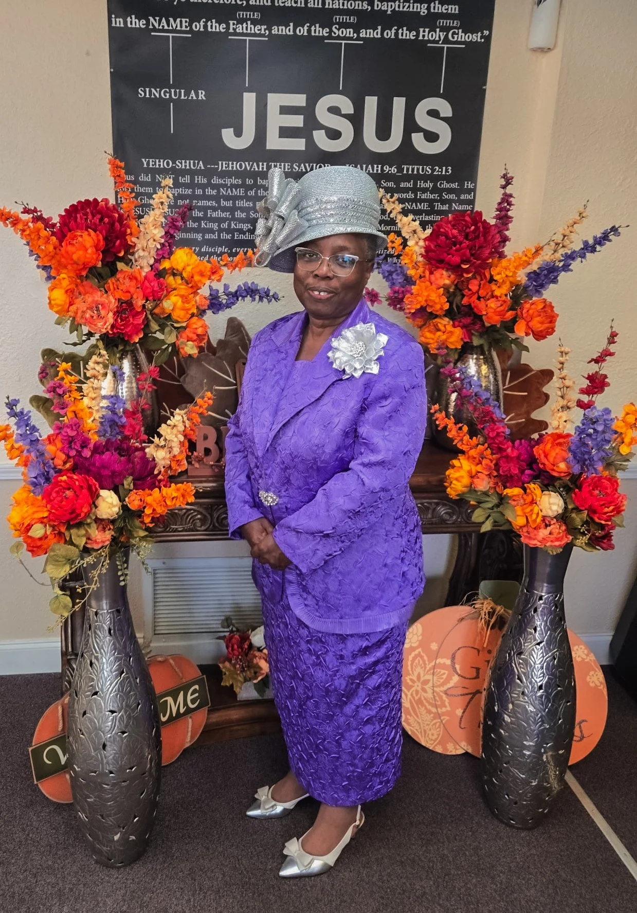 A woman dressed in a purple lace dress with a matching jacket, gray hat, and white shoes, stands between two tall vases with colorful artificial flowers. She is in front of a black banner with religious text about Jesus Christ and Christian beliefs. decorations, including pumpkins and a 'Welcome' sign, are on the floor next to her.