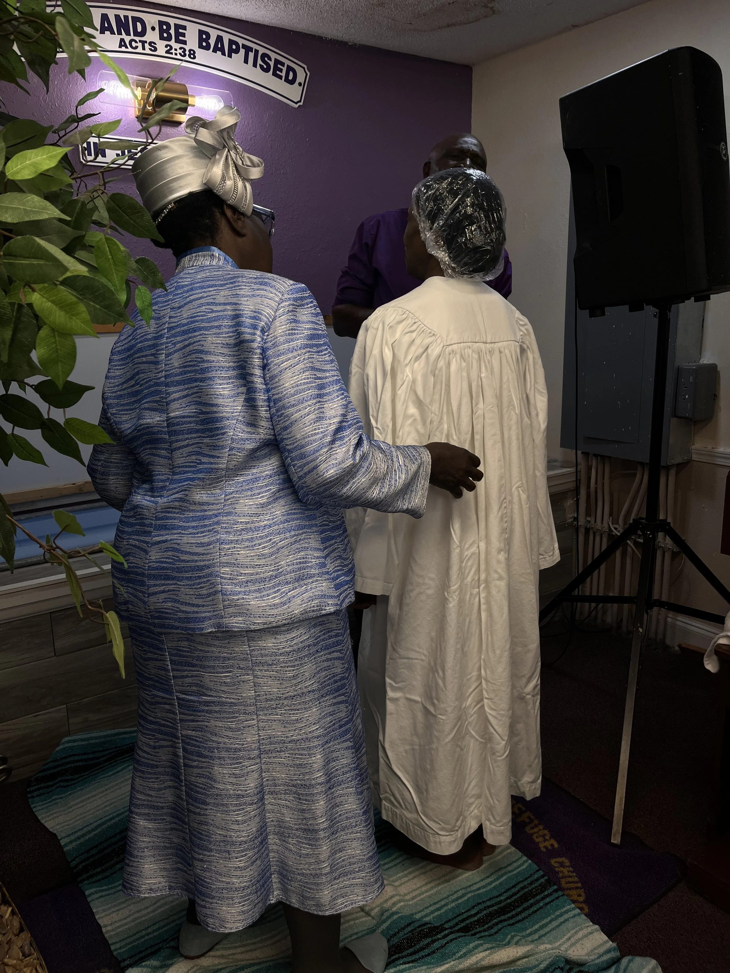 A person wearing a blue patterned robe and a headwrap kneeling on a striped rug during a religious baptism ceremony, with two individuals standing in front of a purple wall with a religious sign reading 'And Be Baptised' from Acts 2:38. One person is
