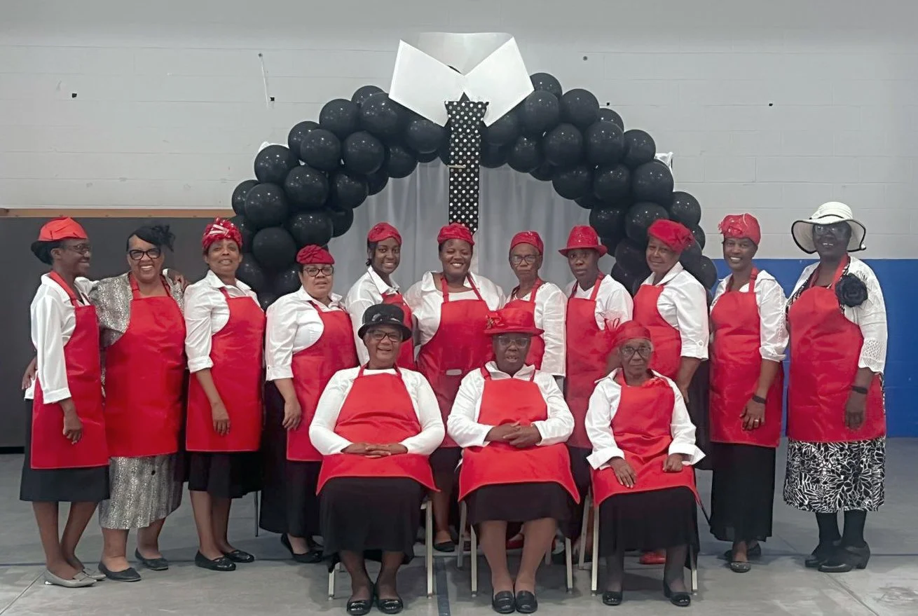 Group of women wearing red aprons and hats, posing in front of a black balloon arch at an indoor event.