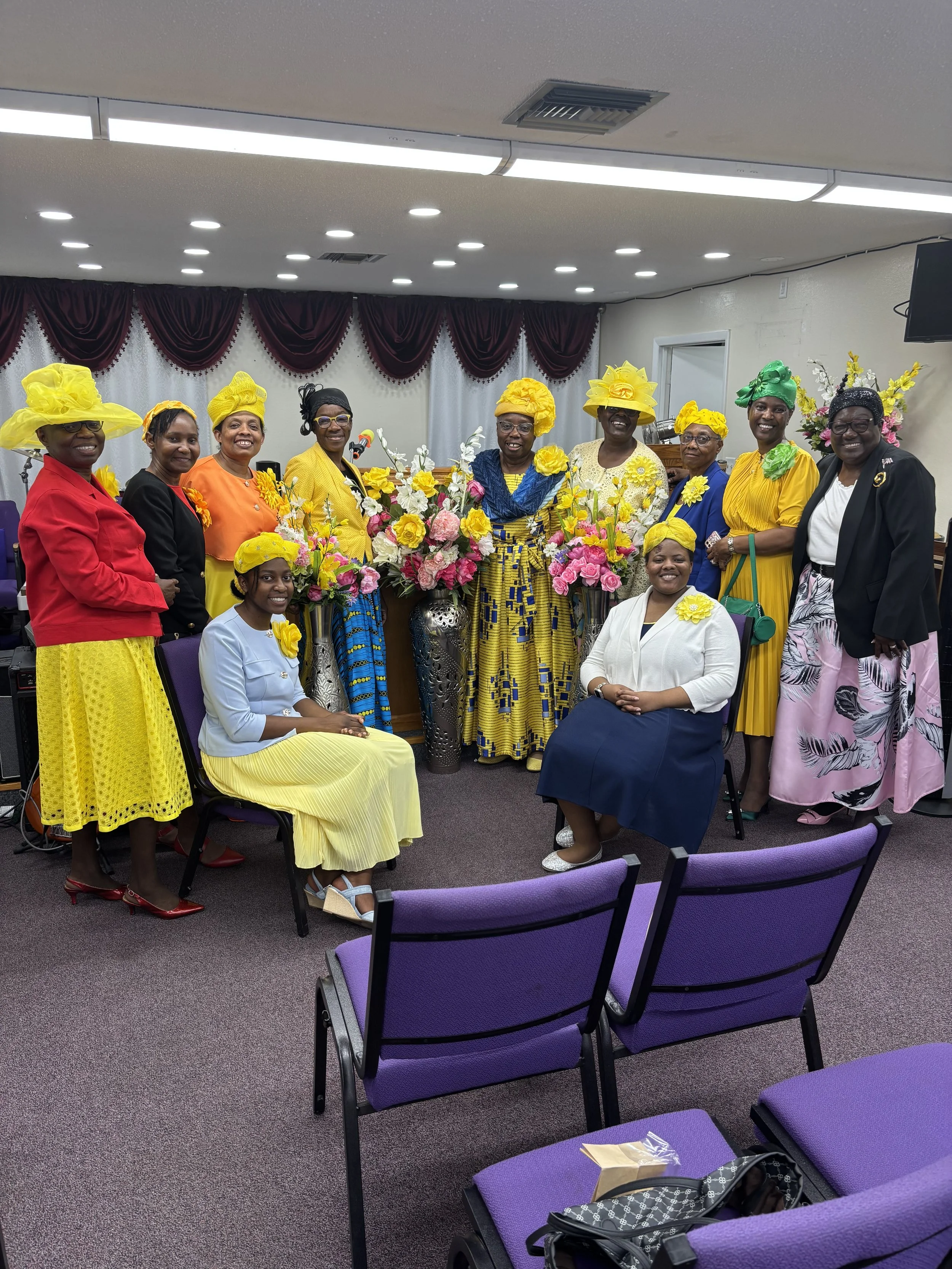Group of women dressed in colorful dresses and large bright yellow hats, gathered in a room decorated with floral arrangements and purple drapes. Some women are standing, and two women are seated in chairs with purple upholstery, one in a yellow skir