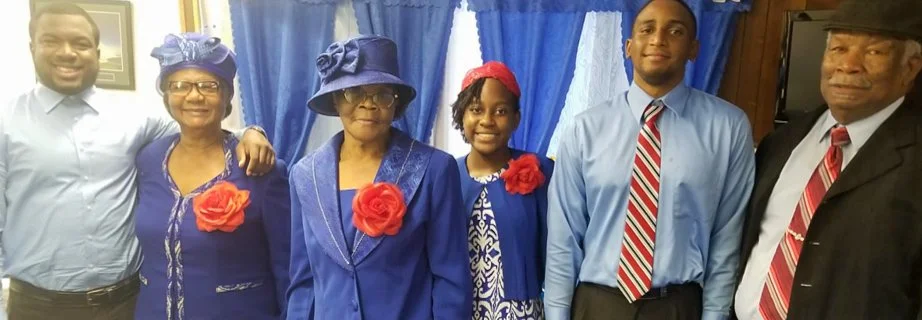 Family group photo with six people, all dressed in formal blue attire, some wearing hats and red flower accessories, standing in front of a blue and white curtain.