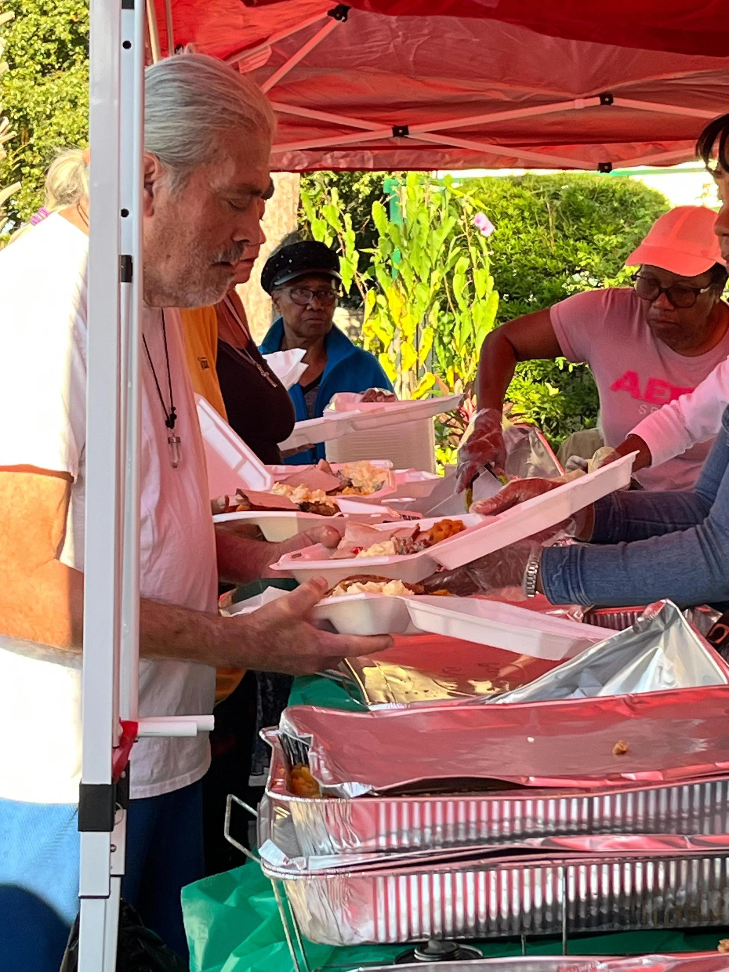 People serving food at an outdoor food bank with a red canopy, green trees, and a blue sky in the background.