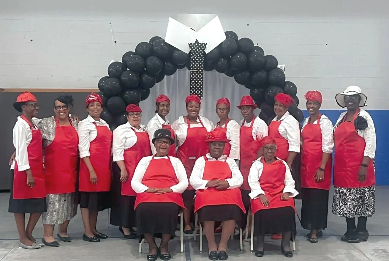 Group of 15 women wearing white shirts, red aprons, and red headscarves, posing in front of a black balloon arch decorated with a white shirt and tie theme. Some women are seated, others standing, in an indoor setting.