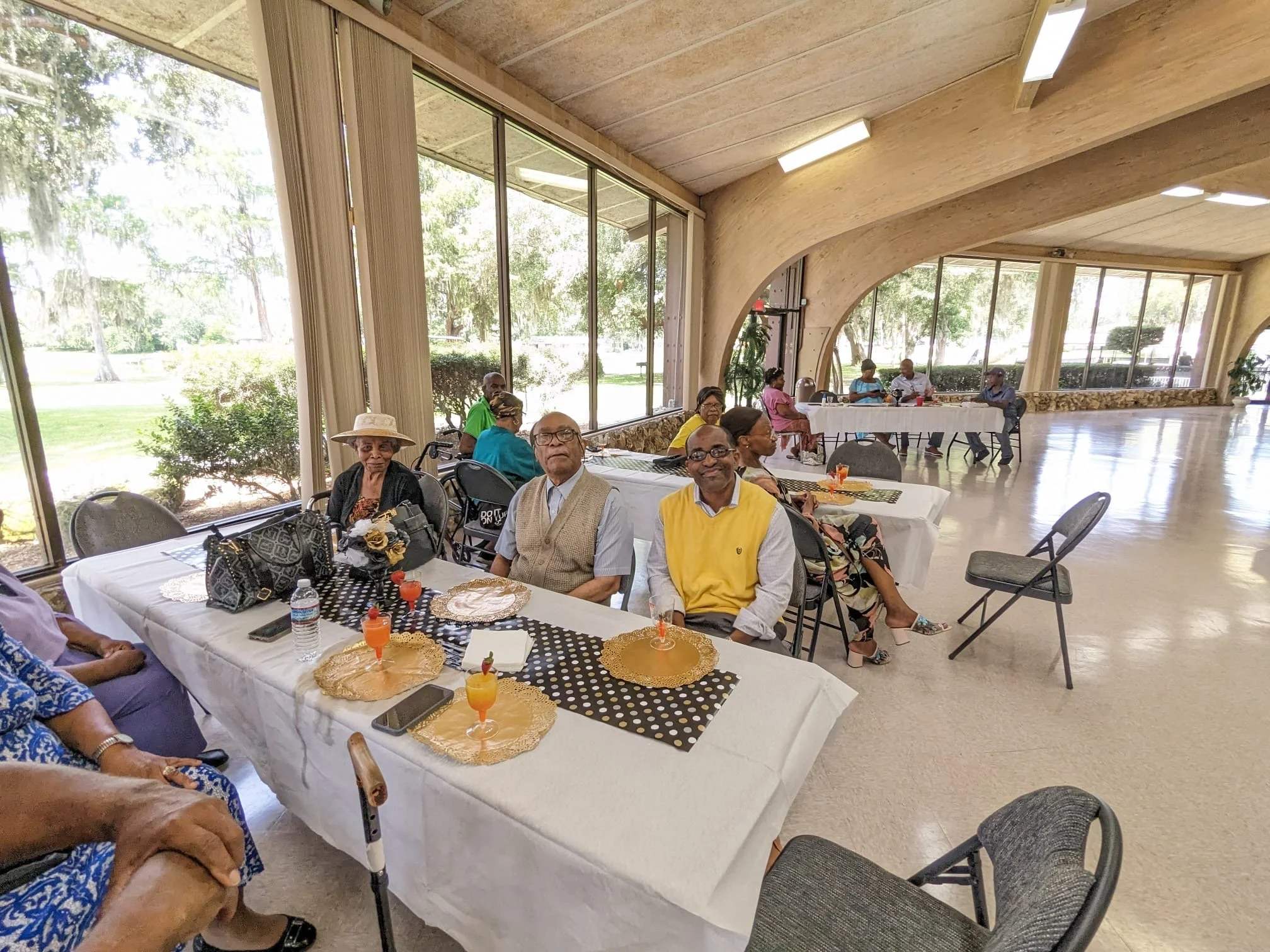 People sitting at tables with refreshments in a spacious room with large windows showing outdoor greenery.