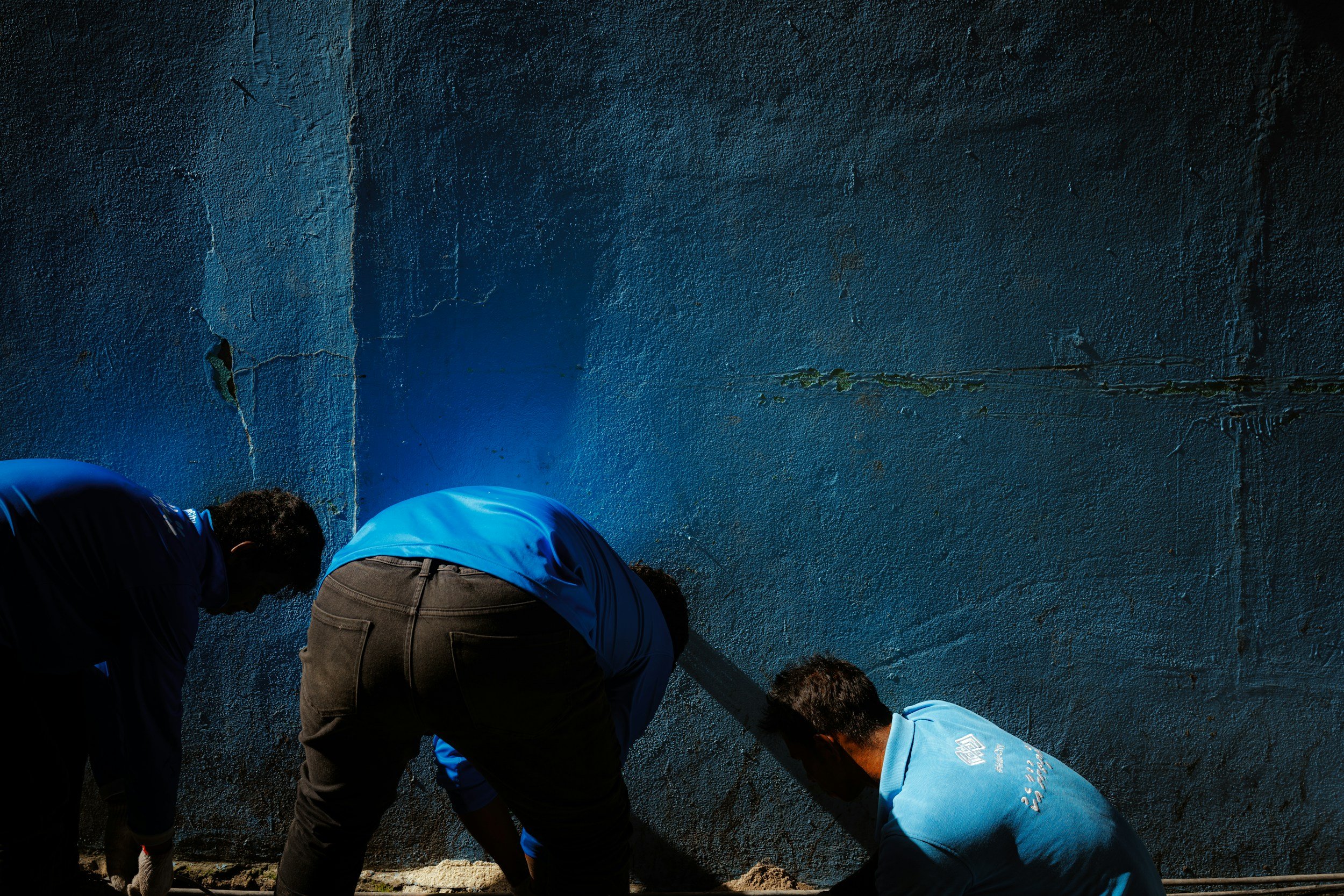 People gathered closely together, leaning forward, against a textured blue wall with peeling paint, wearing casual clothing.