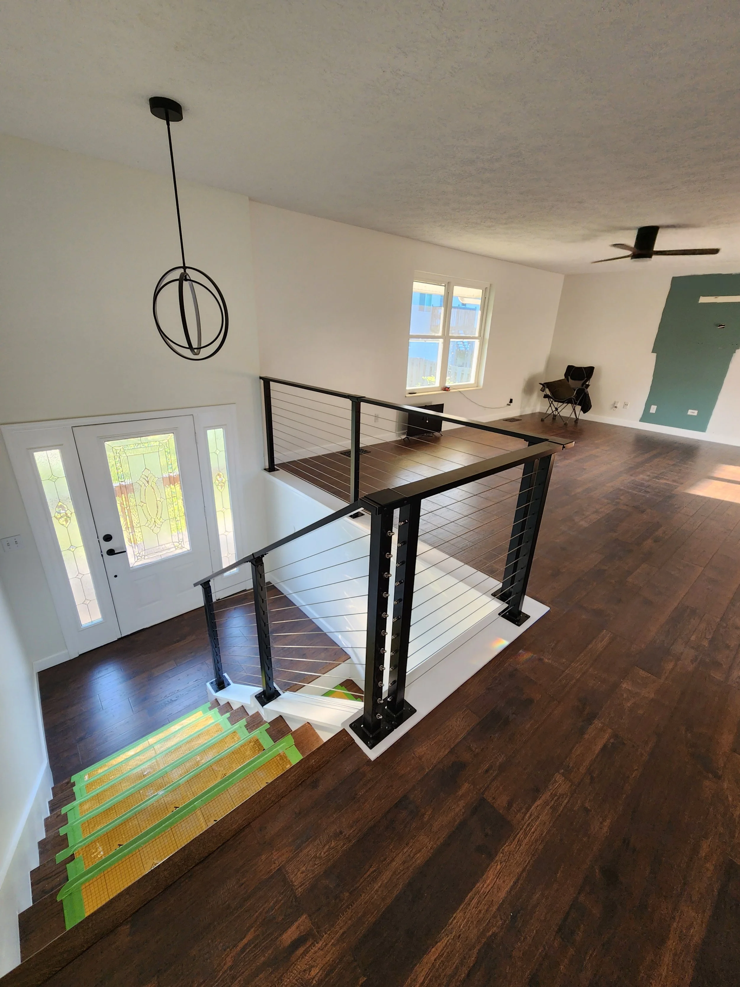 Interior of a modern house with a staircase, wooden flooring, and an entry door with decorative glass panels. There is a hanging light fixture, a ceiling fan, a window, a chair, and a partially painted wall.