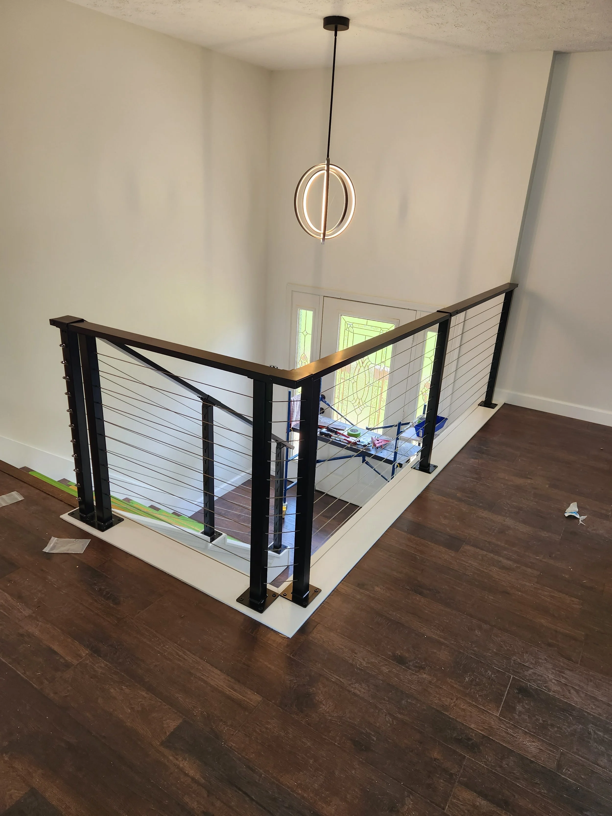 The image shows a modern staircase railing with black metal posts and horizontal wires. The stairwell is illuminated by a circular pendant light hanging from the ceiling. The walls are painted white, and there is a window at the bottom of the stairs with decorative windowpanes. The floor is made of dark wood.