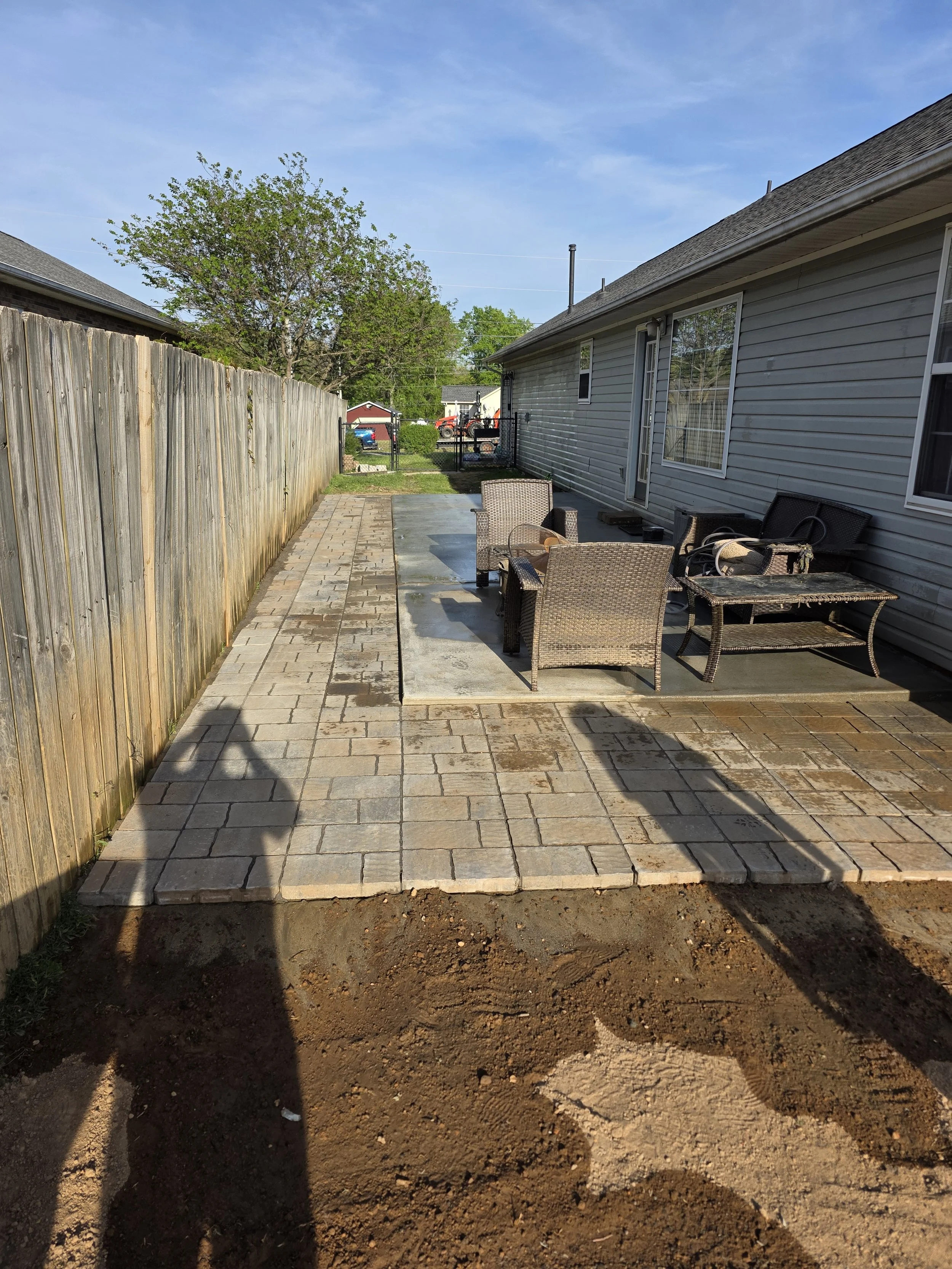 Backyard with a paved patio, outdoor furniture, and a wooden fence, with a house on the right side and trees in the background.