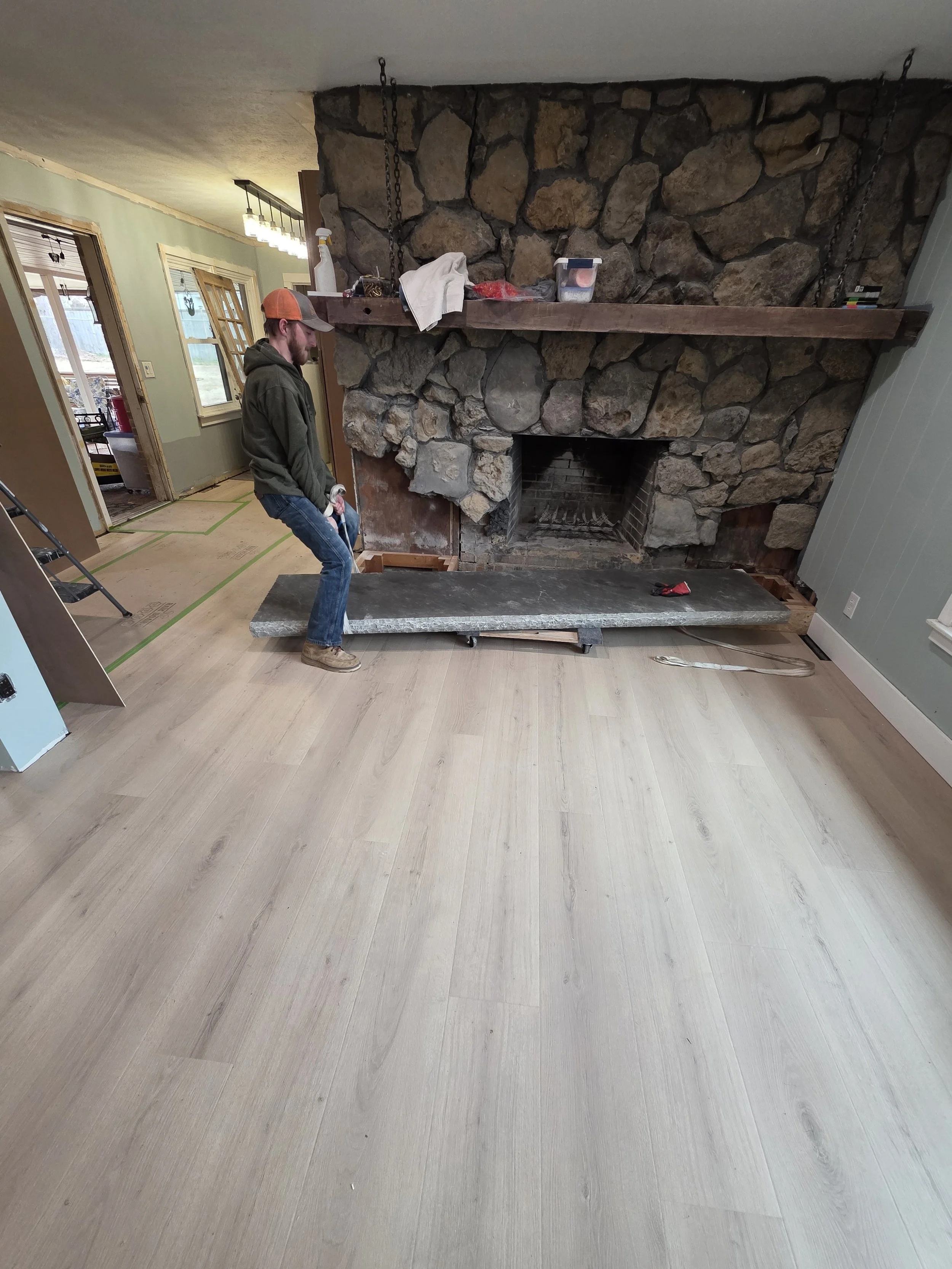 A man in a gray hoodie, jeans, and tan work boots moving a stone hearth in a room with new light wood flooring and a stone fireplace. The room is under renovation, with an open doorway, windows, and construction materials.