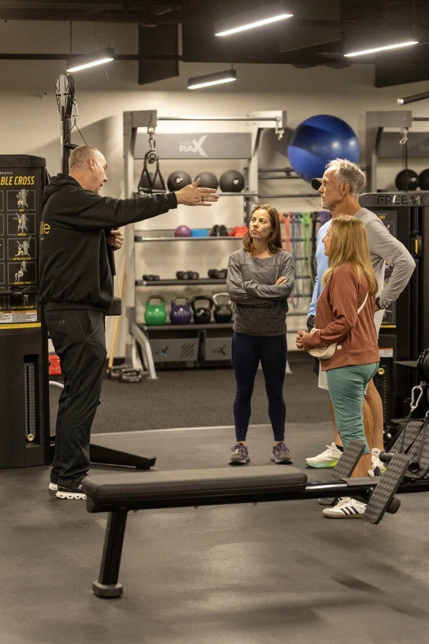 A trainer is explaining an exercise to three adults in a gym. The trainer is gesturing with his hands, and the three adults are listening attentively. The gym has various equipment like kettlebells, medicine balls, and resistance bands in the background.