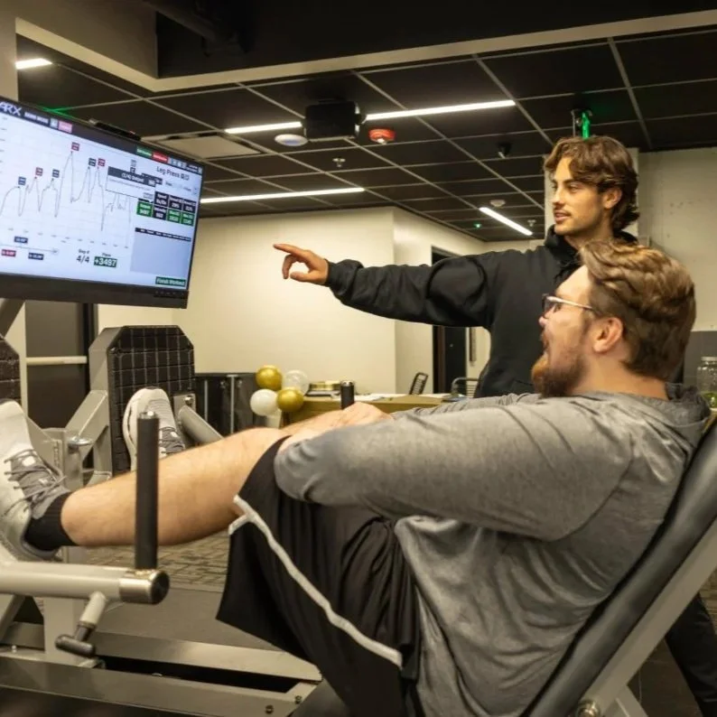 Two men in a gym, one lying on a leg press machine and the other standing, pointing at a screen displaying a workout graph.
