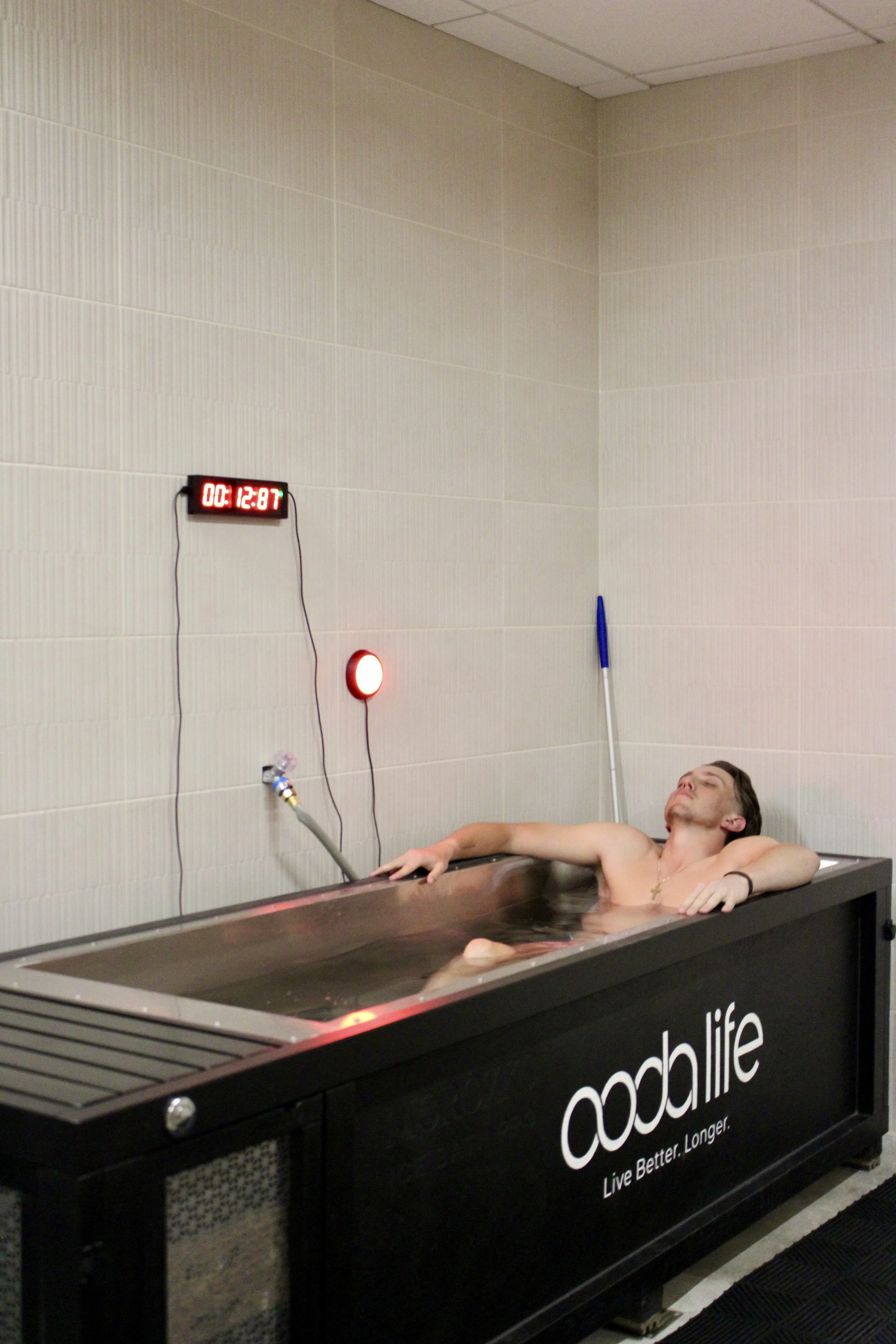 A man is lying in a cooling tub filled with water, with his arms resting on the edge. A digital clock on the wall behind him displays 00:12:87. There is a blue and white item, possibly a tool or medical instrument, leaning against the wall.