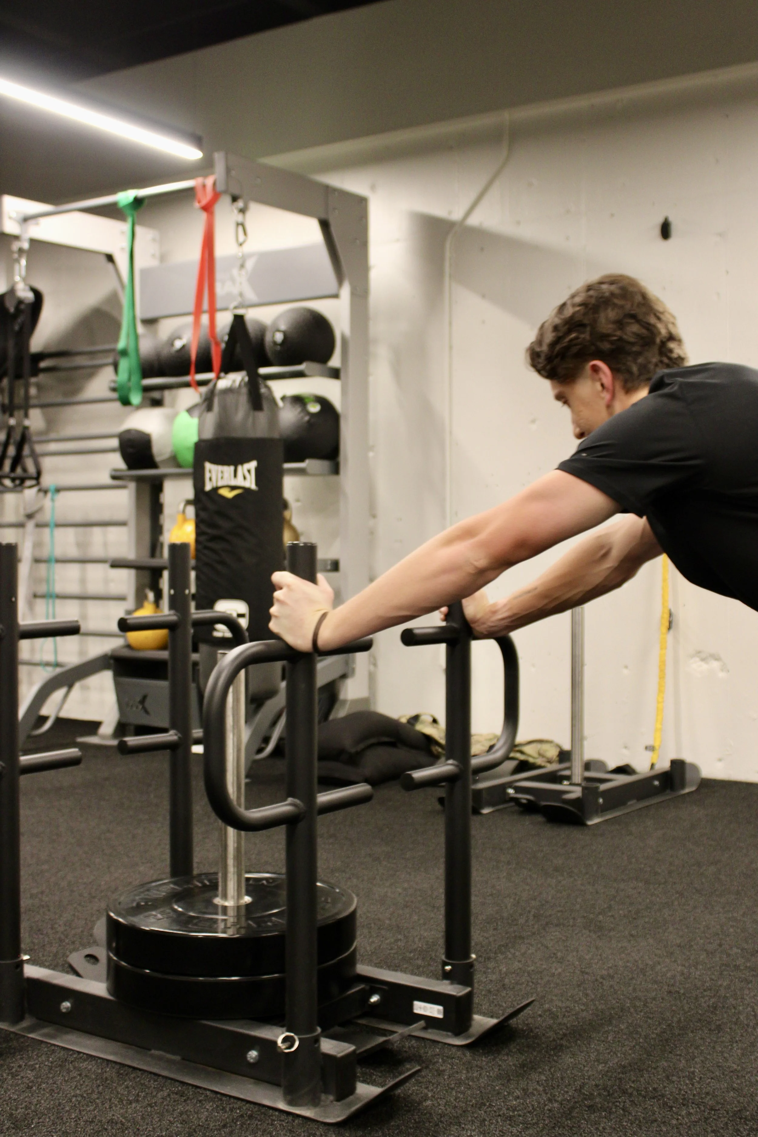 A man doing a stretching exercise in a gym with workout equipment such as a boxing bag, kettlebells, and resistance bands in the background.